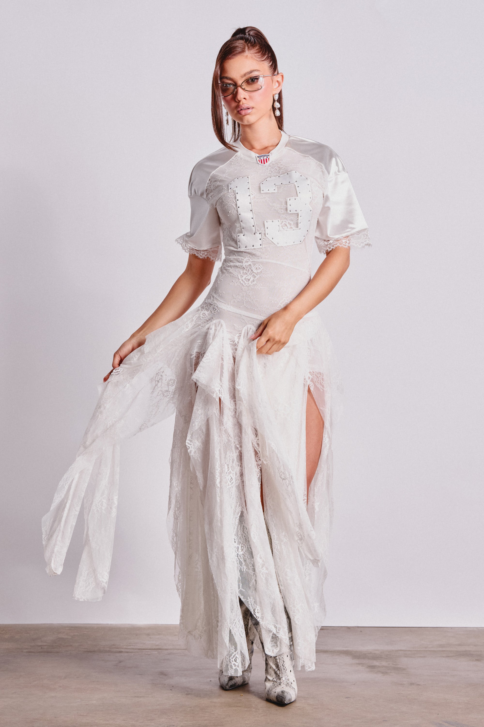 A woman models the KICK OFF LACE JERSEY MAXI DRESS IN WHITE, featuring a sequined sports jersey top with number 13 and a sheer lace skirt. She accessorizes with glasses, a necklace, and patterned boots against a plain background. 1