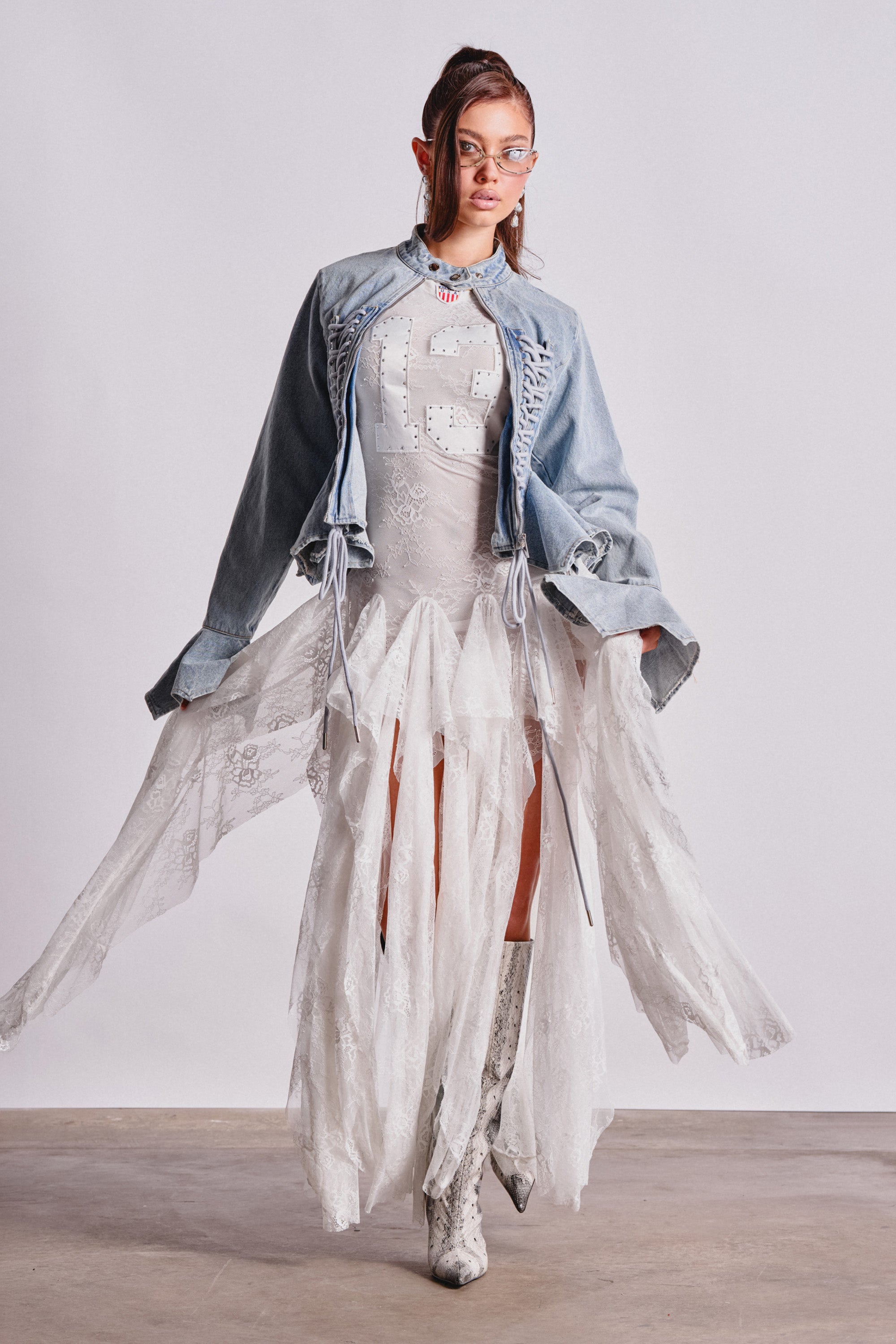 A woman models the KICK OFF LACE JERSEY MAXI DRESS IN WHITE under a light blue denim jacket, styled with glasses and patterned knee-high boots for a chic, sporty look against a plain light background. 2