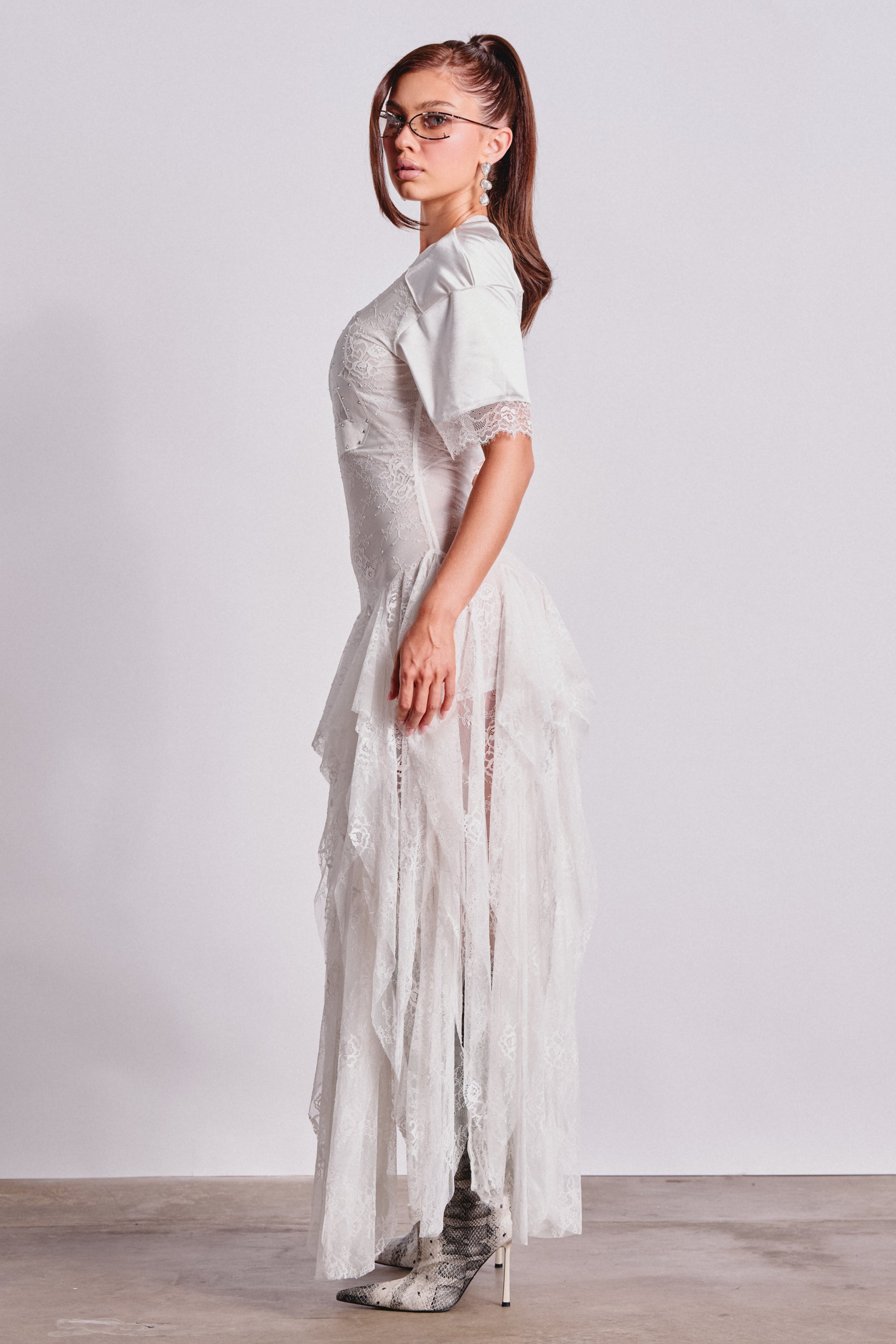 A woman with long, straight brown hair in glasses stands in profile against a plain background, wearing the KICK OFF LACE JERSEY MAXI DRESS IN WHITE over a white t-shirt, paired with patterned ankle boots. 3