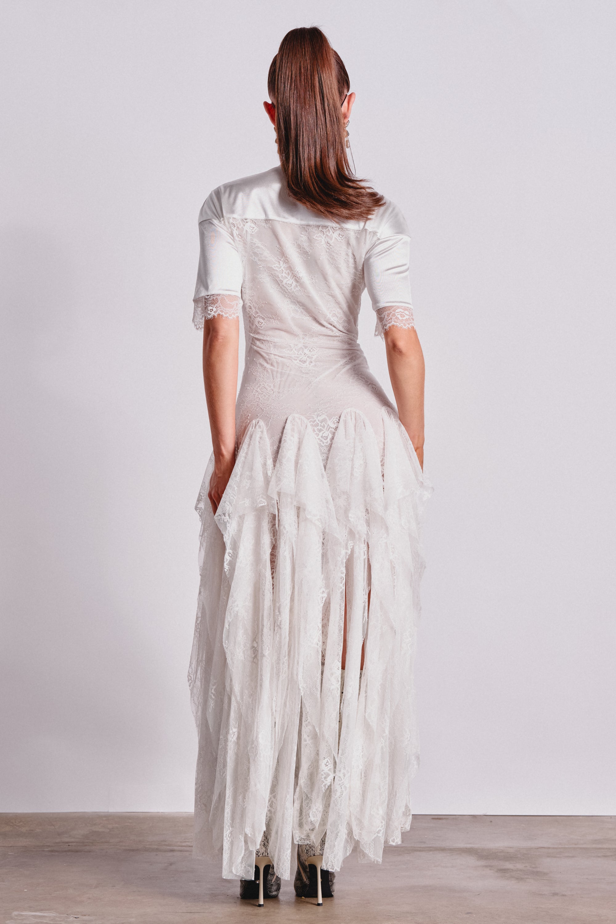 A person stands facing away in the KICK OFF LACE JERSEY MAXI DRESS IN WHITE, featuring layered ruffles and short sleeves. Their straight brown hair is in a high ponytail against a plain, light-colored background. 4