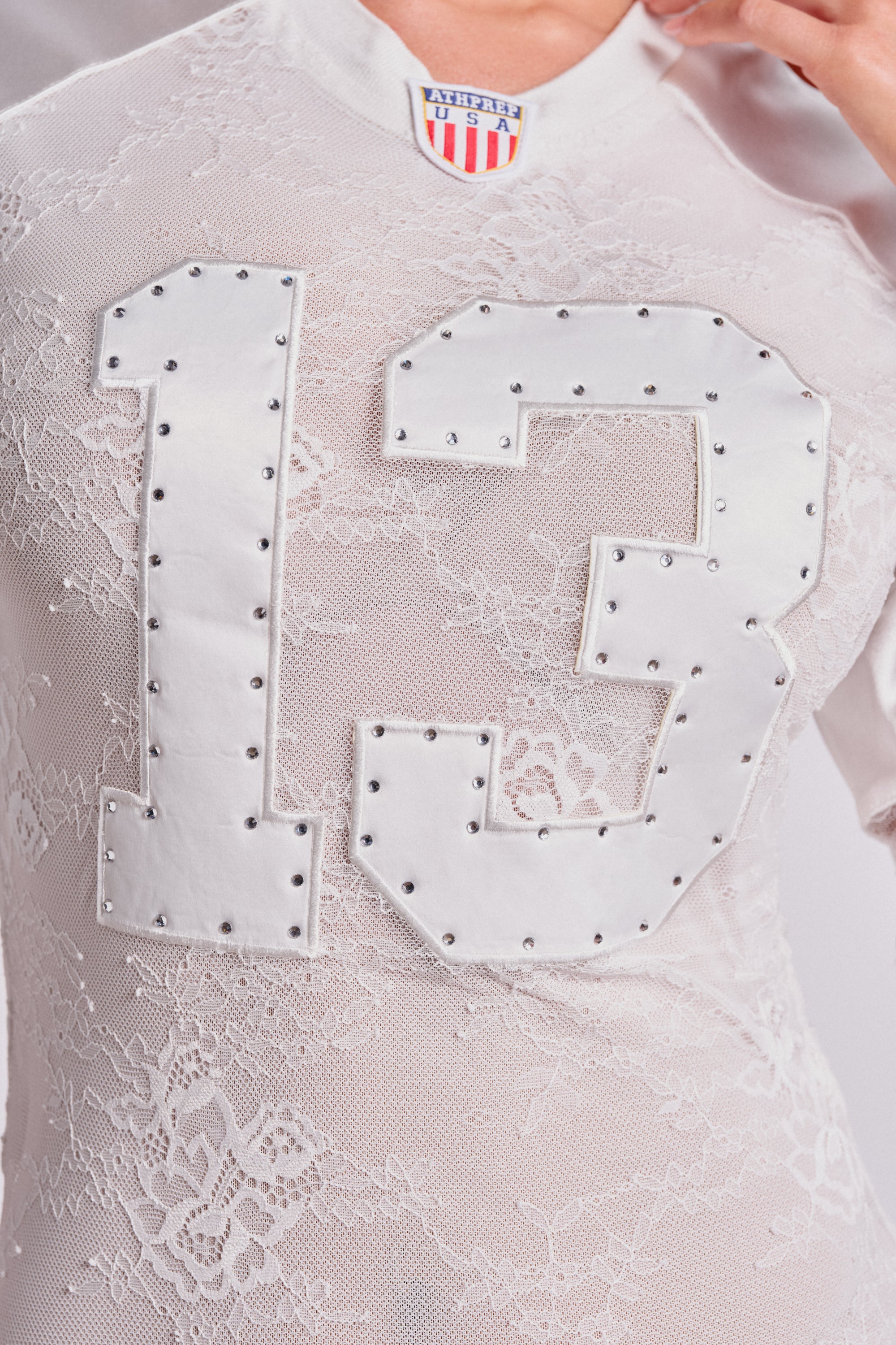 A close-up of a person wearing the KICK OFF LACE JERSEY MAXI DRESS IN WHITE, featuring sporty styling with large white number 13 and silver studs on the front, plus a small American flag tag at the neckline. 5