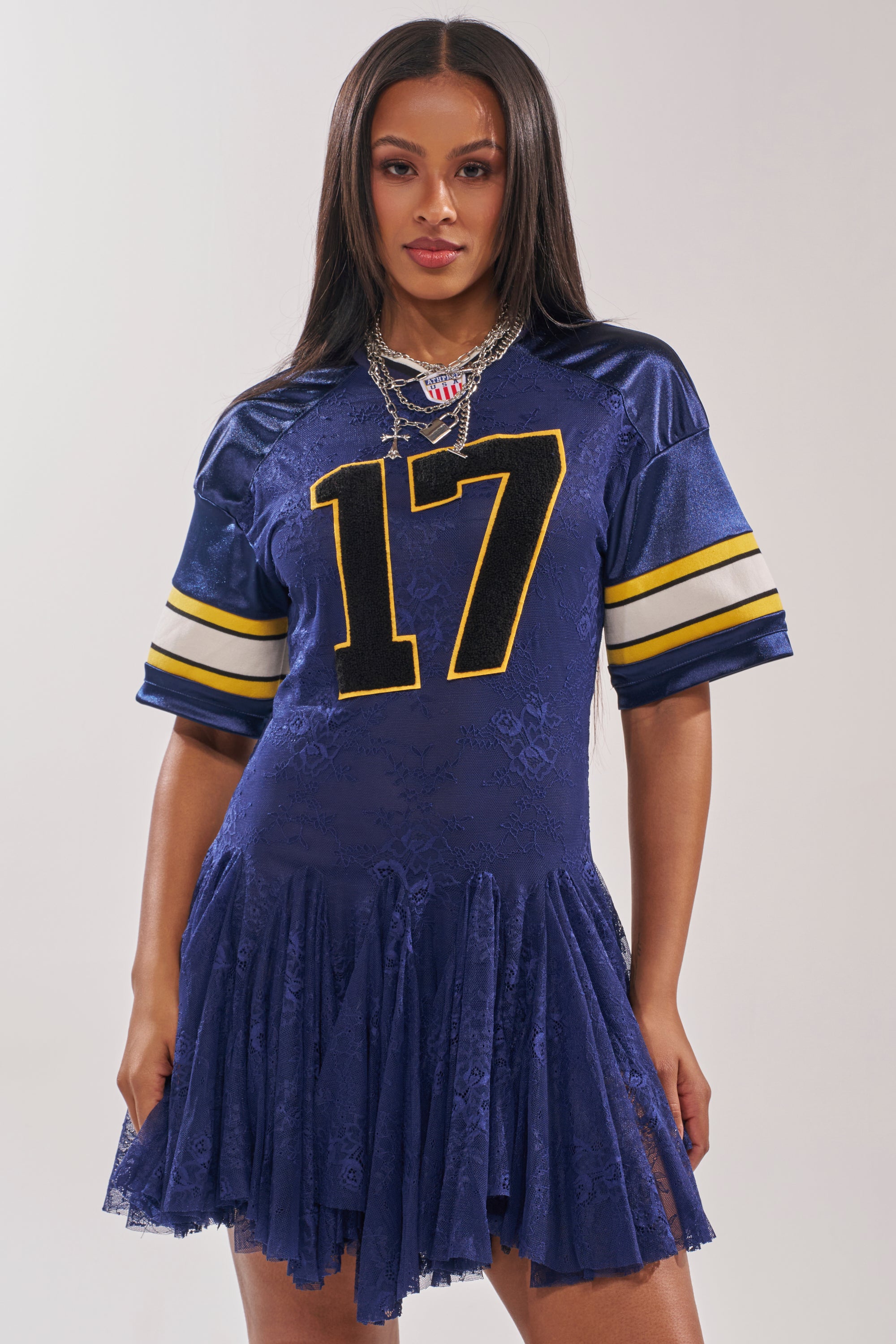 A woman wears the VARSITY BLUES LACE MINI DRESS—a navy football jersey with yellow stripes and the number 17, styled with a flared lace skirt. She poses against a plain light background, her long straight hair complemented by layered necklaces. 1