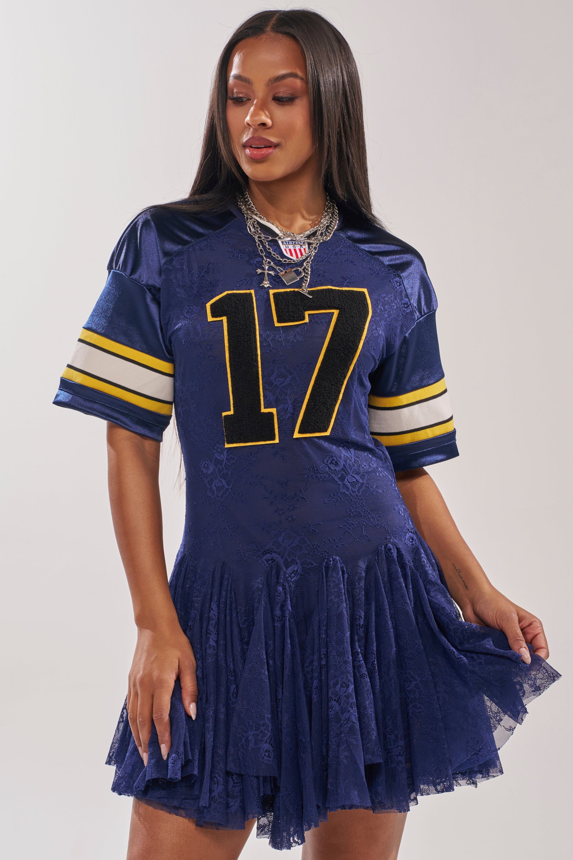 Wearing the VARSITY BLUES LACE MINI DRESS—a navy lace mini dress styled with a navy football jersey featuring yellow number 17 and stripes—she pairs long straight hair and layered silver necklaces for a chic game day look. 2