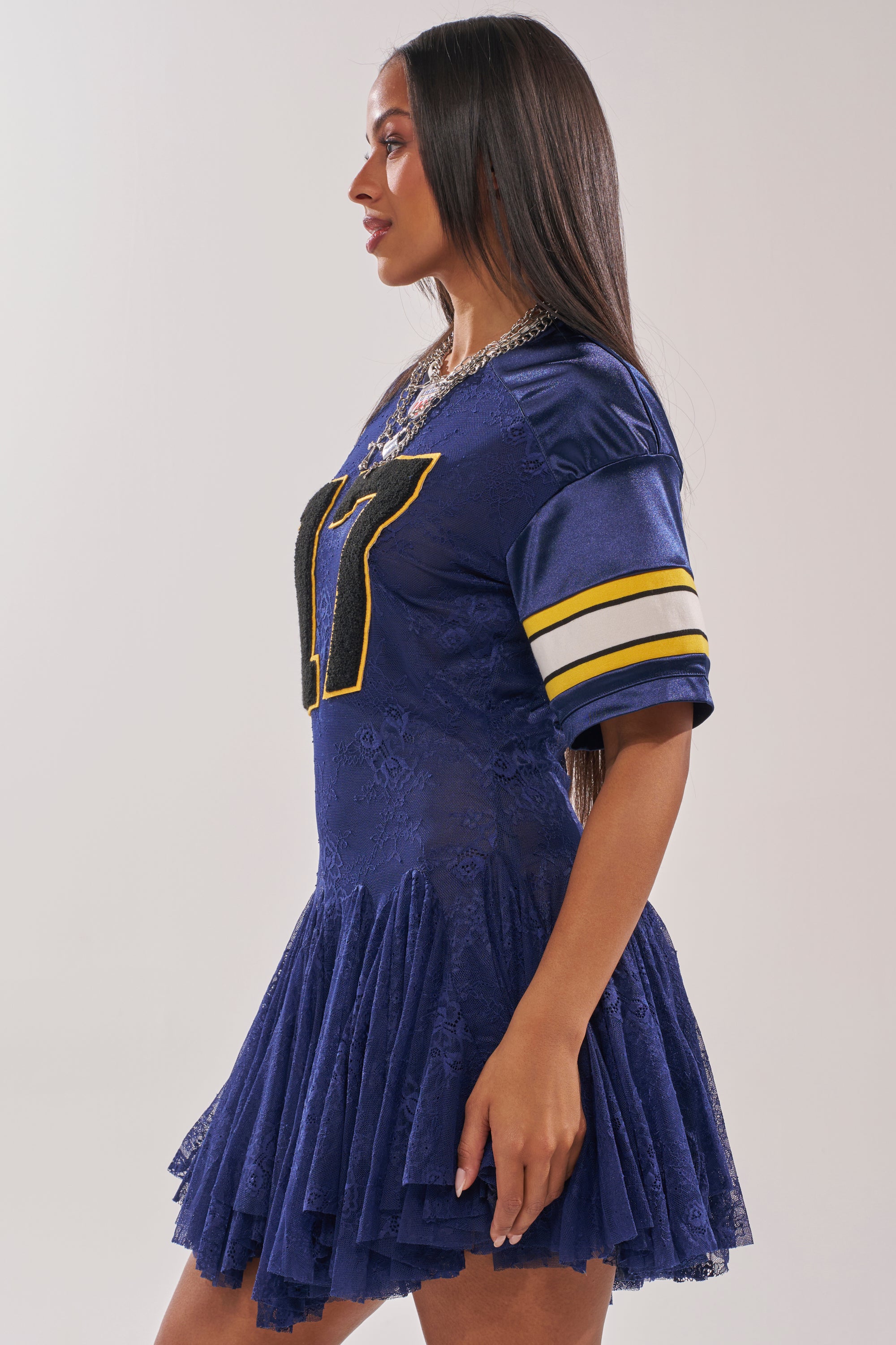 A woman in profile models the VARSITY BLUES LACE MINI DRESS, featuring navy lace, sporty yellow and white sleeve stripes, a bold "17" on the front, and her long straight hair worn down. 3