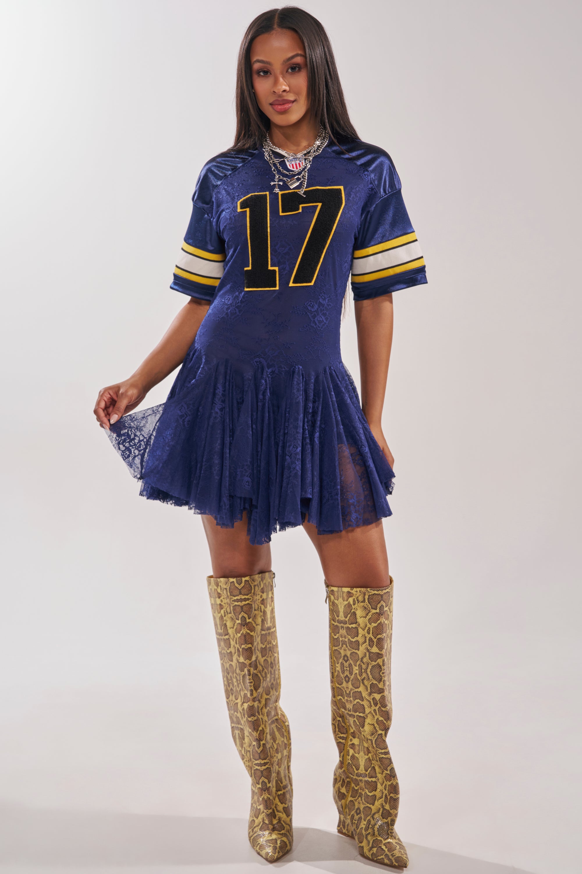 A woman models the VARSITY BLUES LACE MINI DRESS—a sporty blue mini dress with yellow stripes and the number 17—styled with tall yellow snakeskin pointed-toe boots and bold jewelry against a plain background. 5