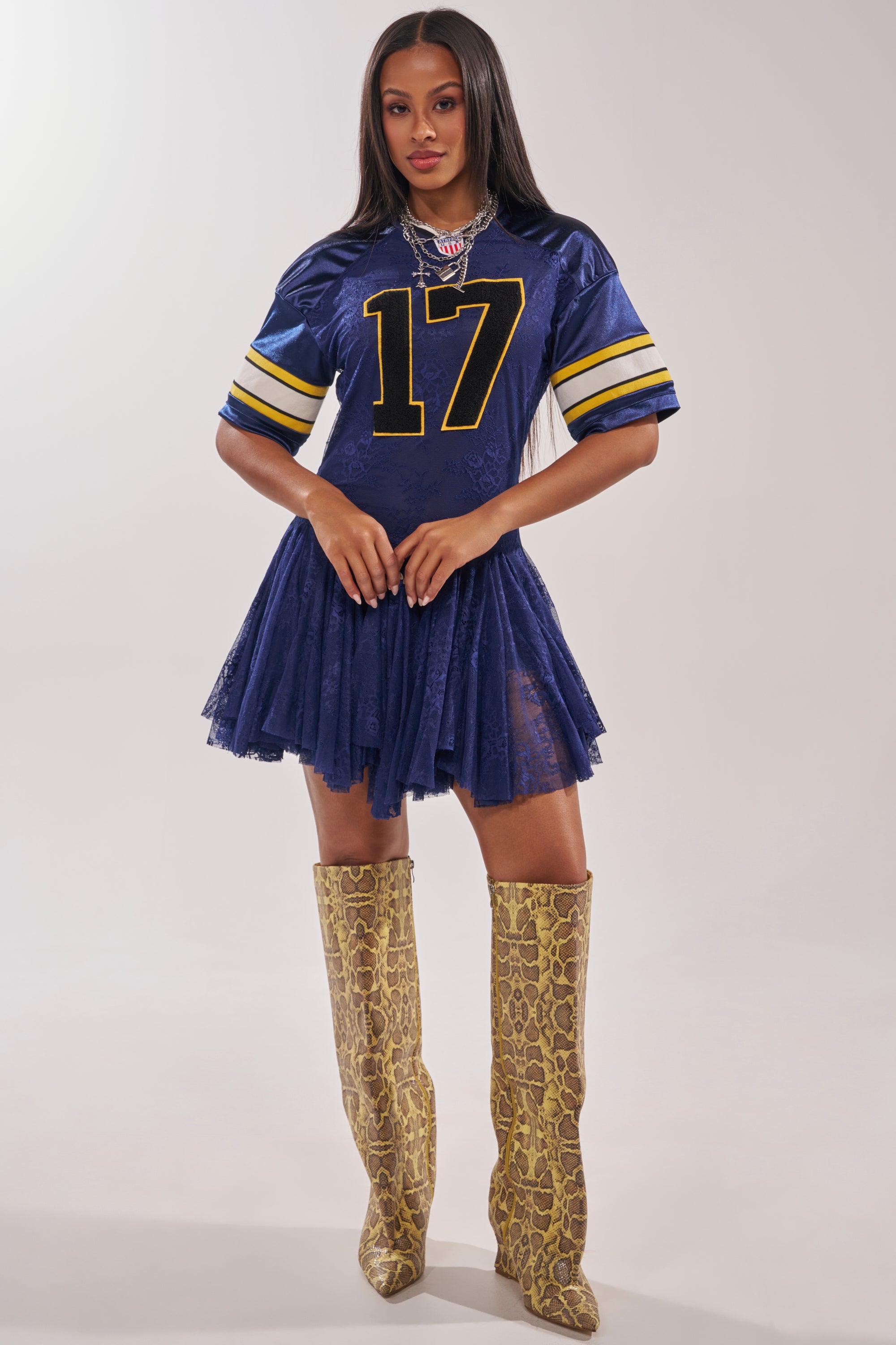 A woman poses with her hands clasped in front, wearing the VARSITY BLUES LACE MINI DRESS—a blue sporty mini dress with yellow stripes and number 17—paired with tall tan snakeskin boots. She has long straight hair. 6