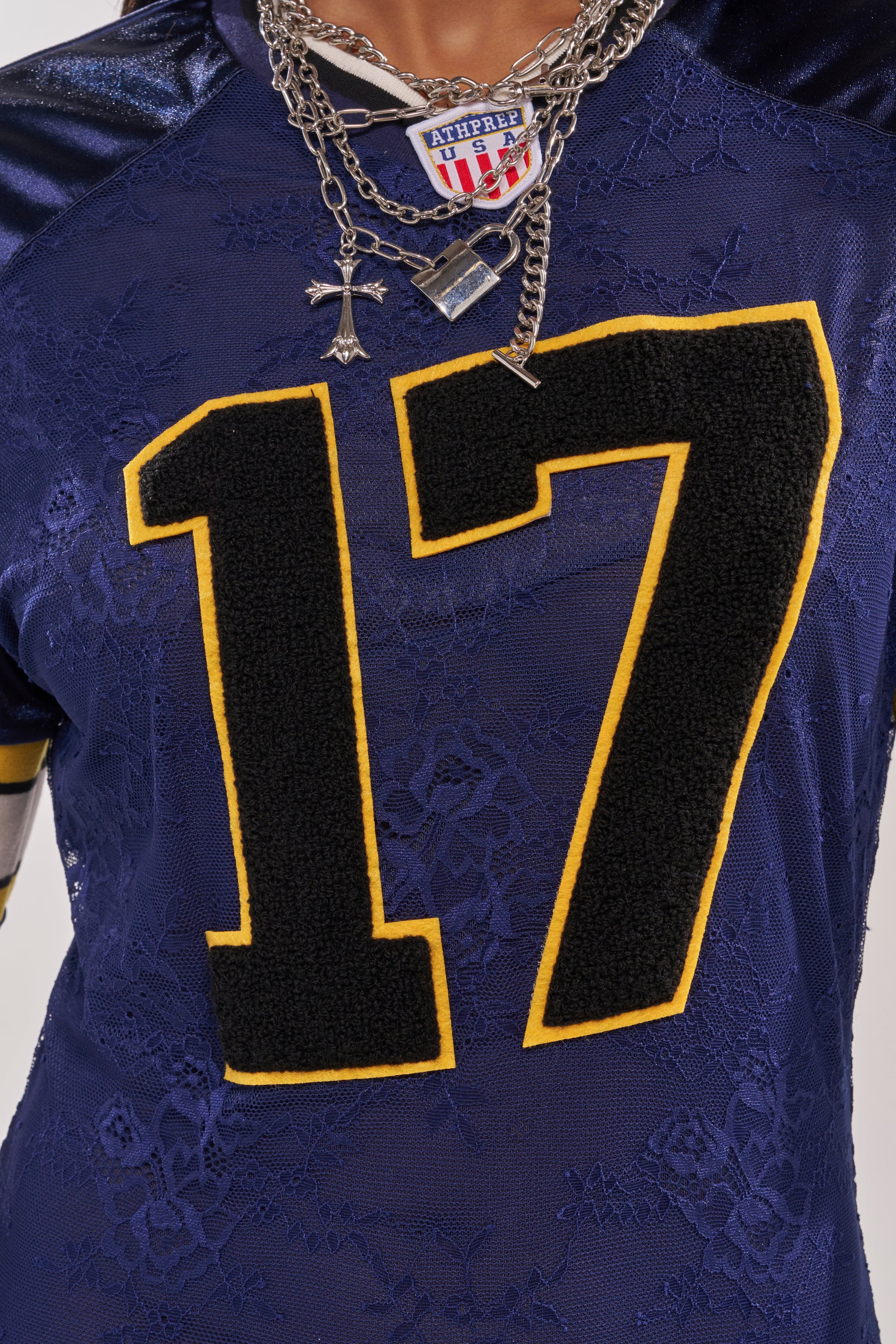 A person wears the VARSITY BLUES LACE MINI DRESS, a sporty dark blue mini dress with bold yellow and black number 17, styled with layered silver necklaces featuring a cross and football-themed pendant. 7