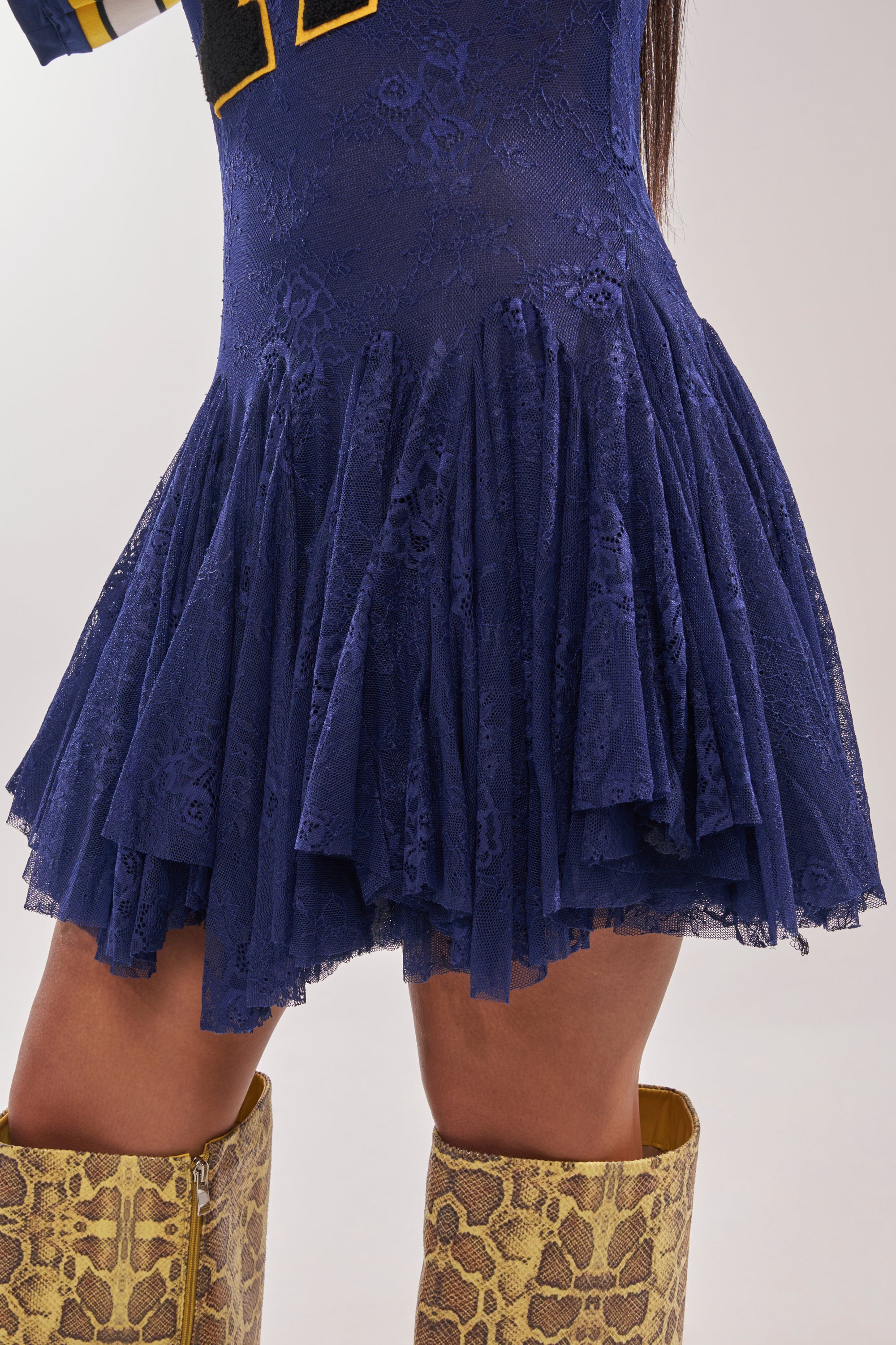 A close-up of a person wearing the VARSITY BLUES LACE MINI DRESS with a flared skirt and tall yellow snakeskin boots, showing only the lower half of the dress and boots against a plain background. 8