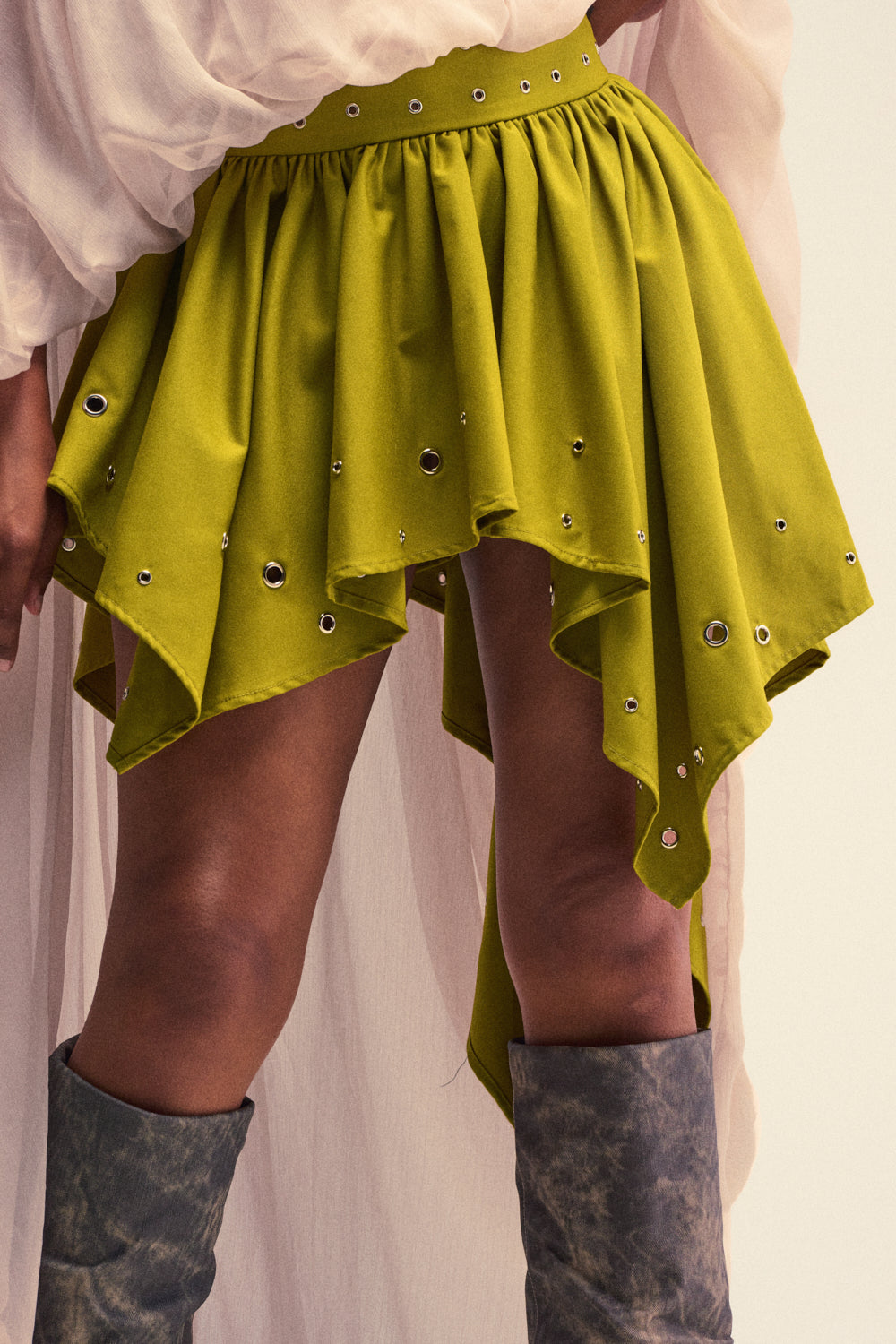 A person wears the YOUNG AND FREE MINI SKIRT WITH GROMMETS in a vibrant green, styled with a white sheer top and tall, distressed gray boots. The photo is cropped from waist to mid-thigh to highlight the skirt’s silver grommet details—an ideal look for bold festival fashion inspiration.