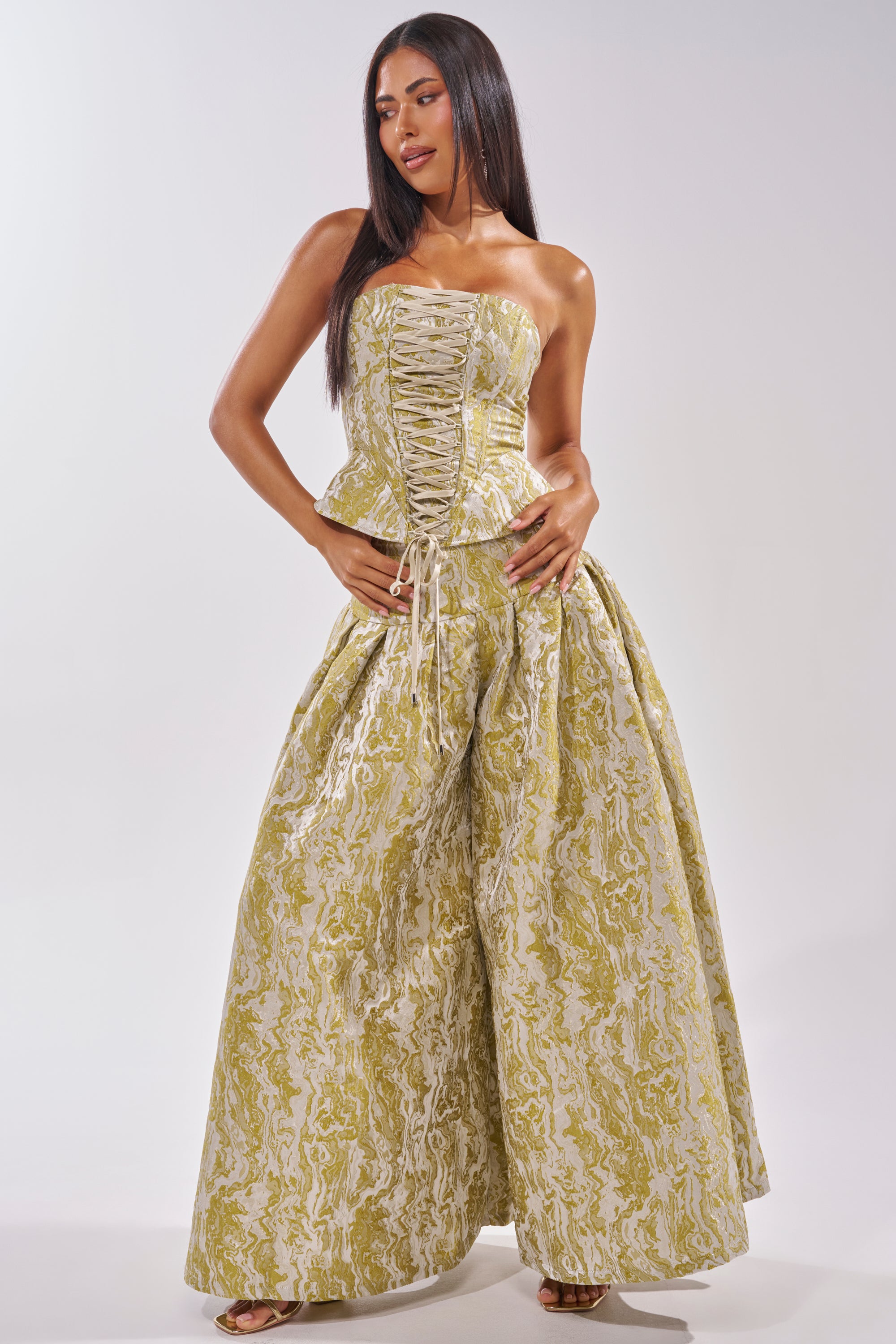 A woman with long straight hair wears a strapless, lace-up corset top and the GET IT FLORAL TROUSER—wide-leg, high-waisted pants with a yellow and white ornate pattern. She stands against a plain light background, looking to the side. 0
