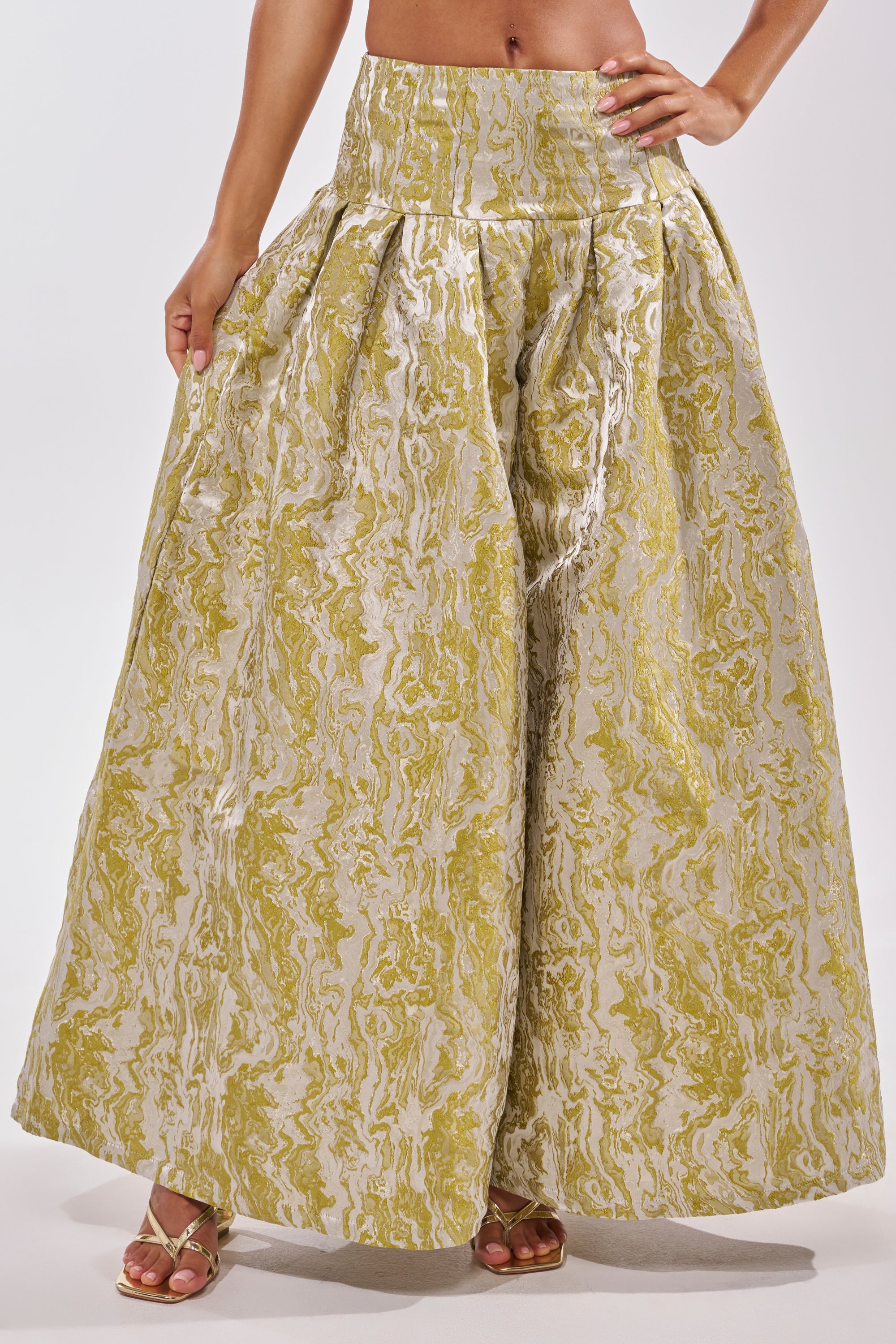 A person wears the GET IT FLORAL TROUSER—wide-legged, high-waisted green brocade pants with a shiny yellow and silver marbled pattern—floor-length for statement brunch style, paired with gold sandals and a confident hand on their hip. 1