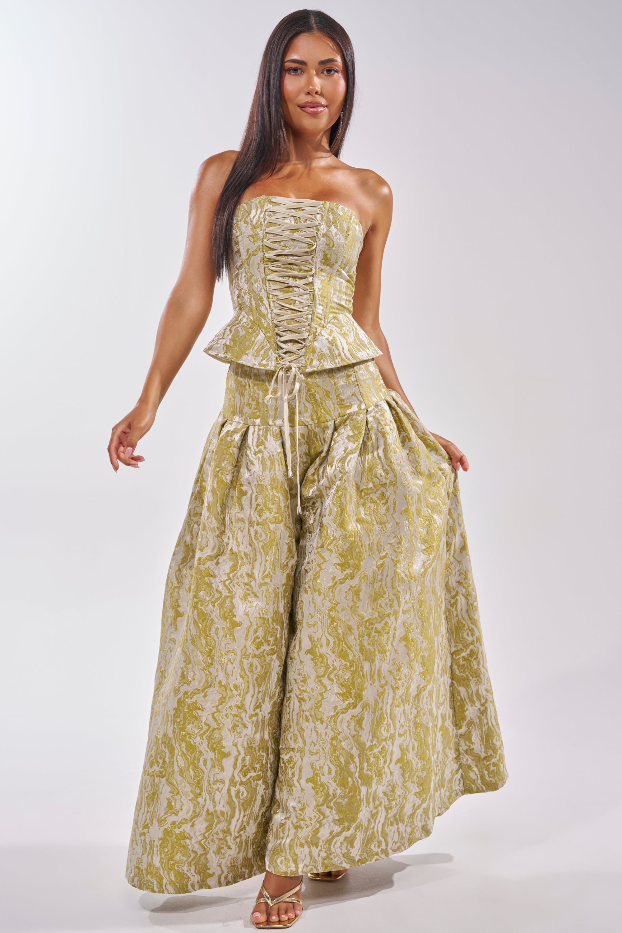 A woman wears the GET IT FLORAL CORSET—strapless and green—with wide-legged, high-waisted yellow and white pants, standing and smiling with one hand holding her skirt for a standout wedding guest look. 5