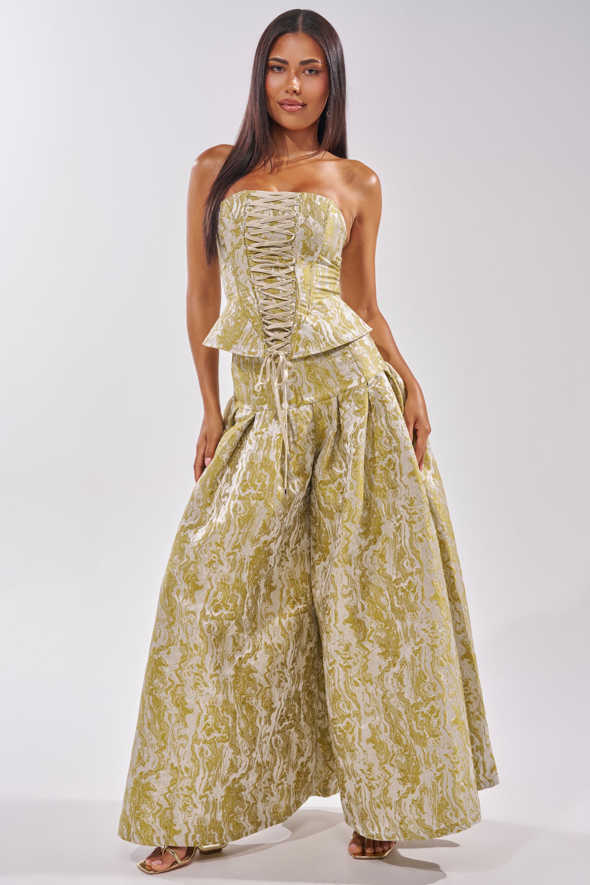 A woman wears the GET IT FLORAL CORSET, a strapless green swirl brocade peplum top, paired with wide-leg, high-waisted pants featuring an intricate green and white pattern—a bold brunch look as she stands facing forward. 7