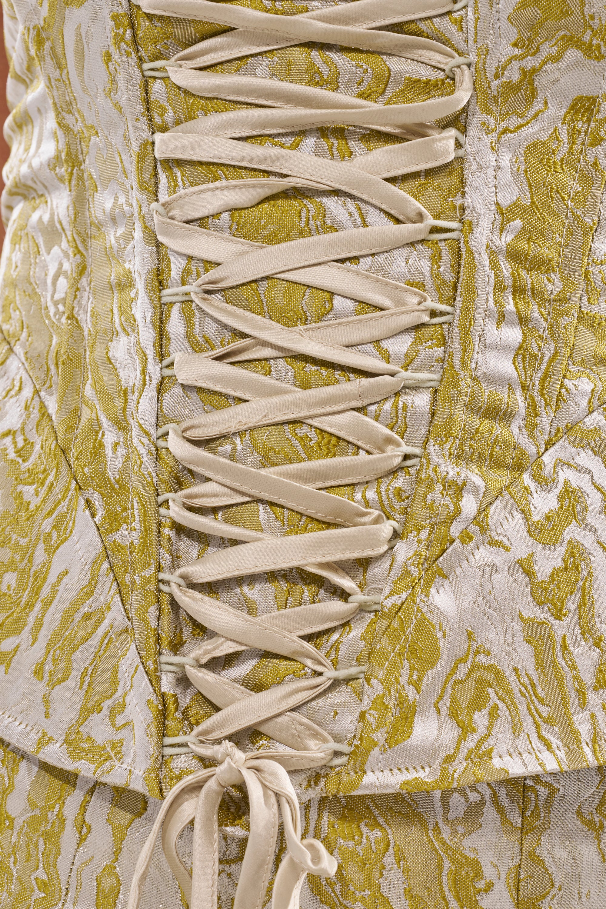 Close-up of the GET IT FLORAL CORSET in luxurious gold and cream brocade with intricate cream laces through metal eyelets. Its textured, wavy pattern makes it a standout choice for brunch or as a bold wedding guest outfit. 8