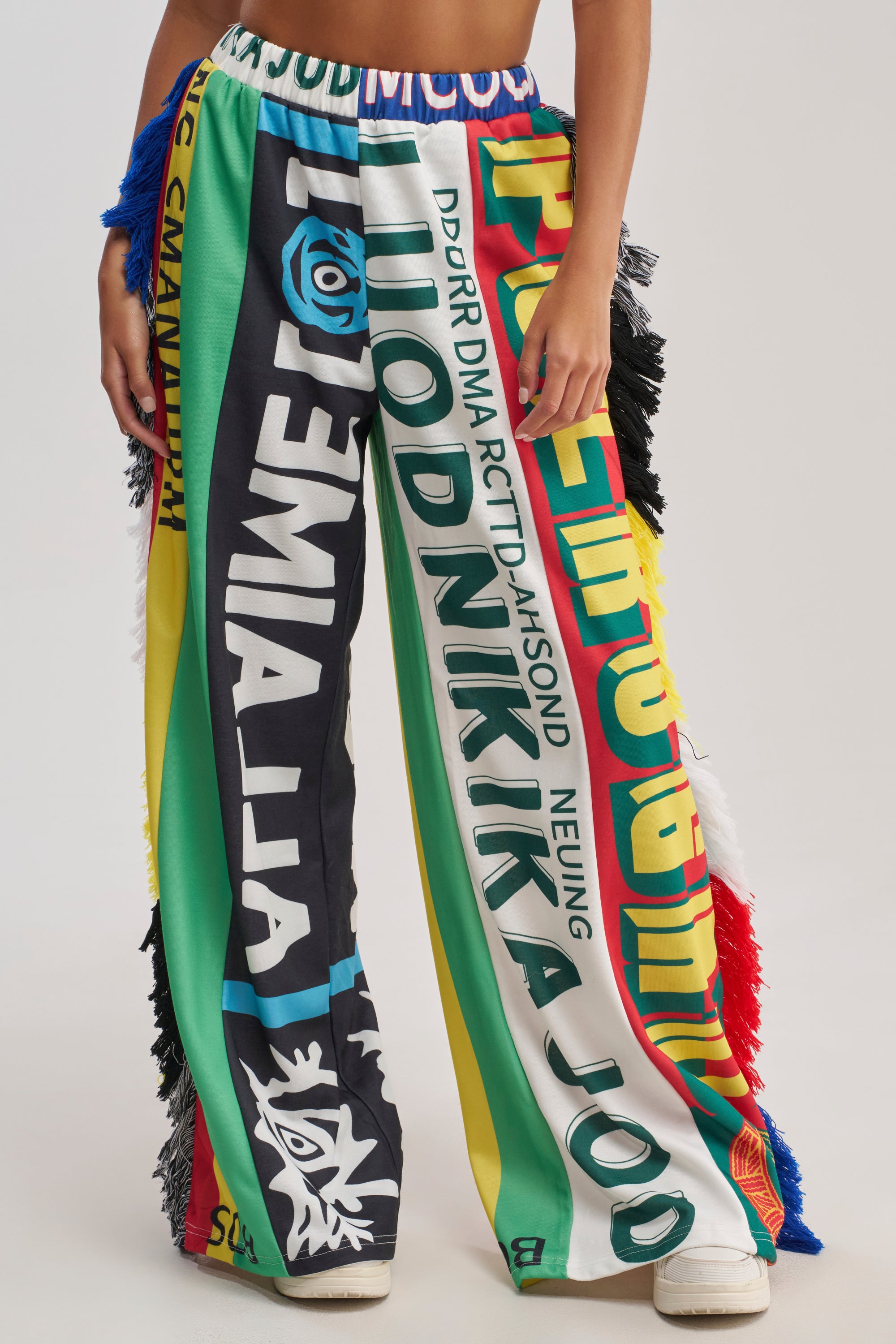 Wearing the MIXED EMOTIONS GRAPHIC JOGGER, featuring colorful wide-leg pants with bold graphic patterns, multicolored text, and fringed sides, paired with white shoes. Only the lower body is visible.