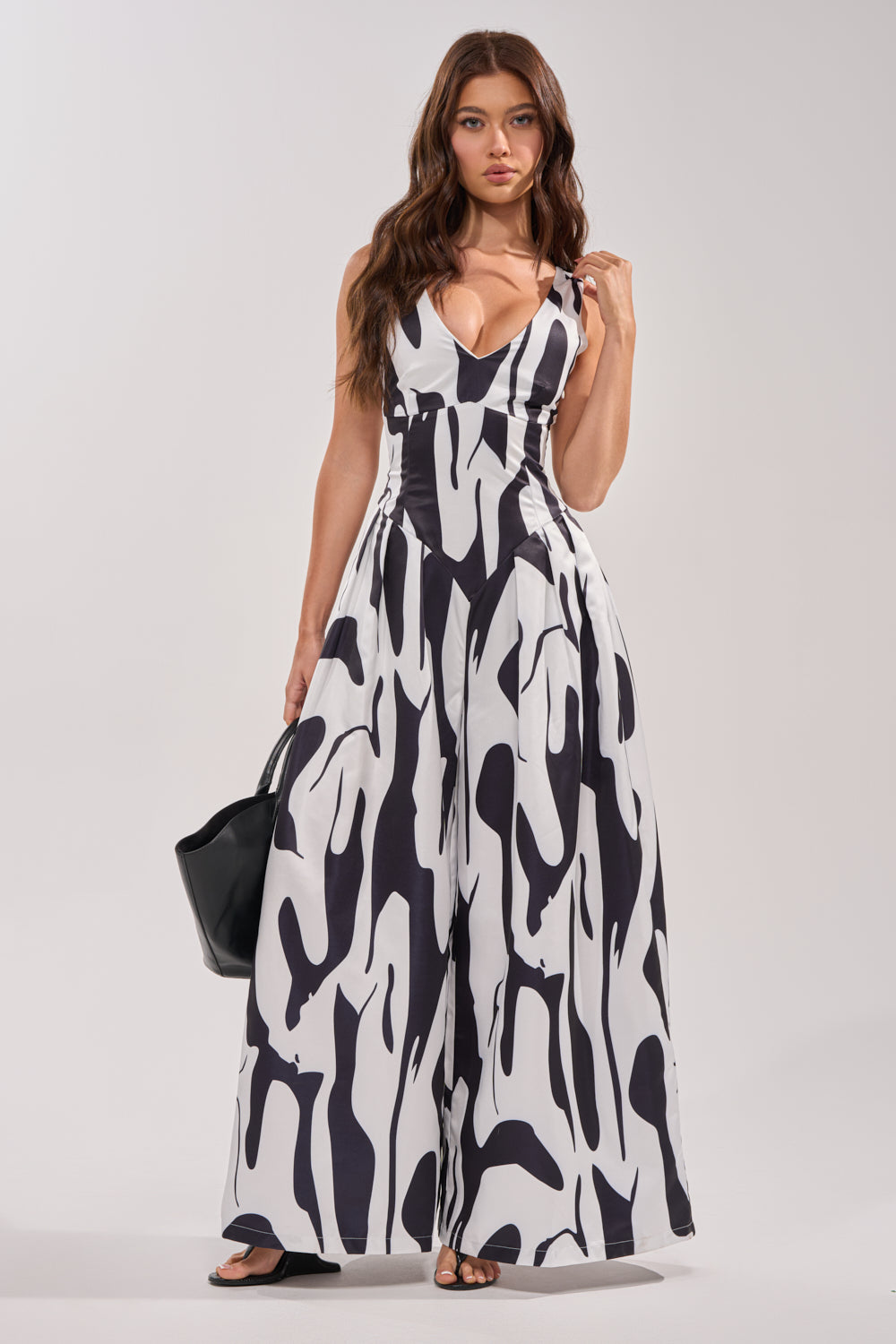 A woman with long brown hair models the READ ALL ABOUT IT POPLIN JUMPSUIT, a sleeveless black and white abstract print wide-leg romper with a deep V-neck, styled with a black handbag against a light gray background. 1