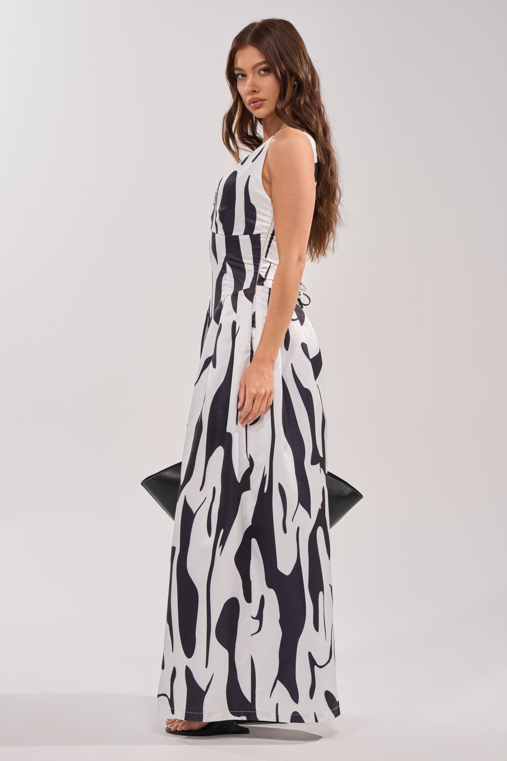 A woman with long brown hair models the READ ALL ABOUT IT POPLIN JUMPSUIT, a black and white piece with bold abstract patterns, standing sideways and holding a black handbag against a plain light background. 2