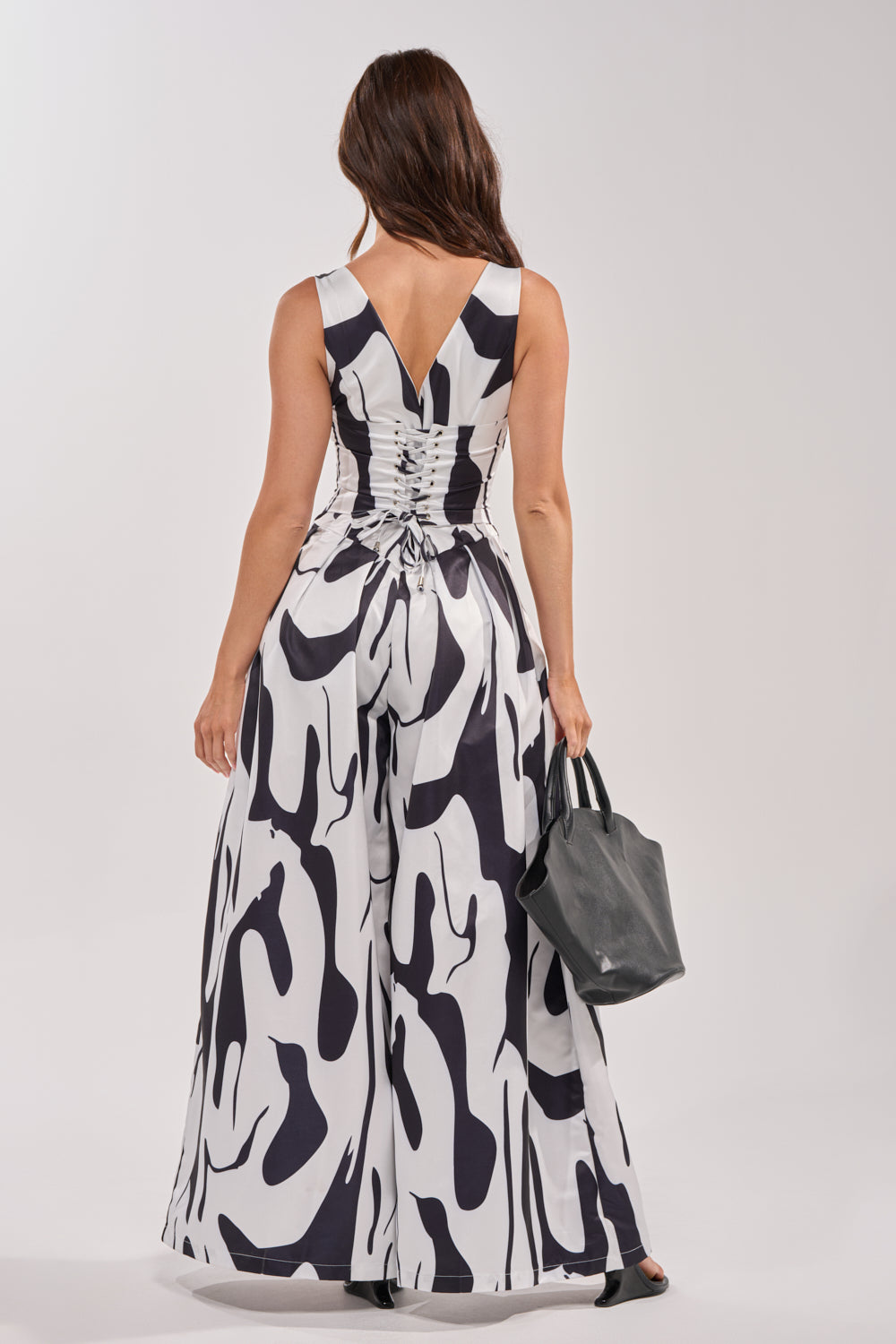 A woman stands facing away, wearing the READ ALL ABOUT IT POPLIN JUMPSUIT—an abstract black and white romper with a V-shaped ruched back detail—while holding a gray handbag. 3