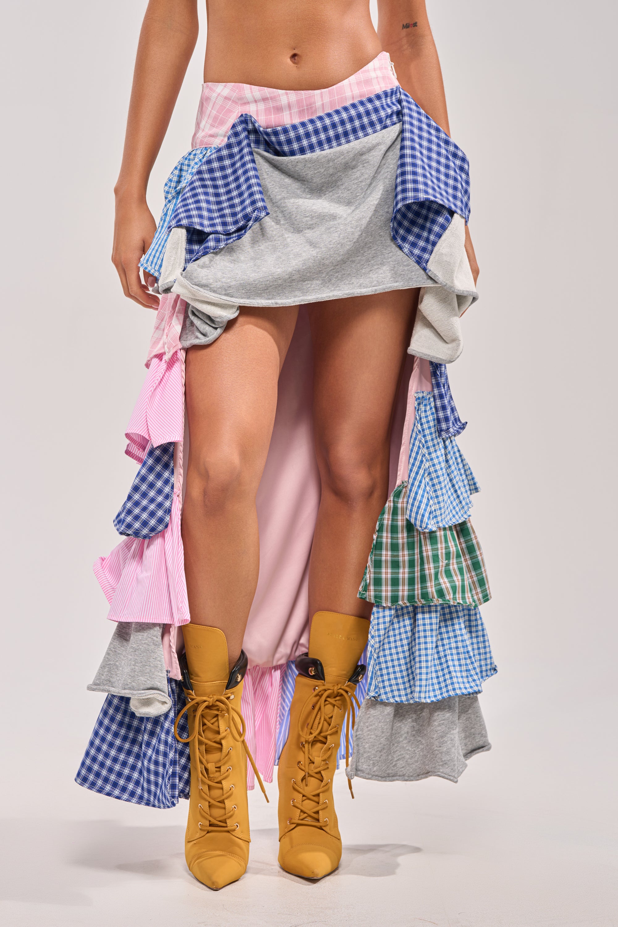 A person wears the MIXED EMOTIONS PLAID HIGH LOW SKIRT with blue, pink, and green checks and plaid ruffles, paired with mustard yellow lace-up boots. The upper body is not visible. 2