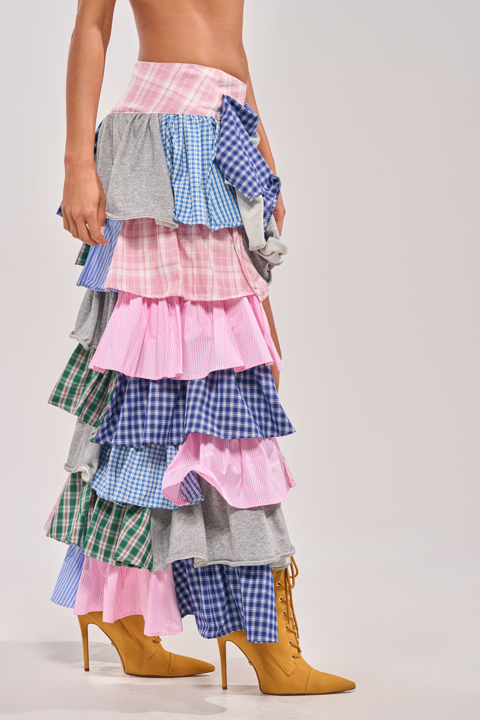 A person wears the MIXED EMOTIONS PLAID HIGH LOW SKIRT—a multi-layered ruffle design in pink, blue, green, and gray—paired with mustard yellow lace-up high-heeled boots. The background is light gray. 4