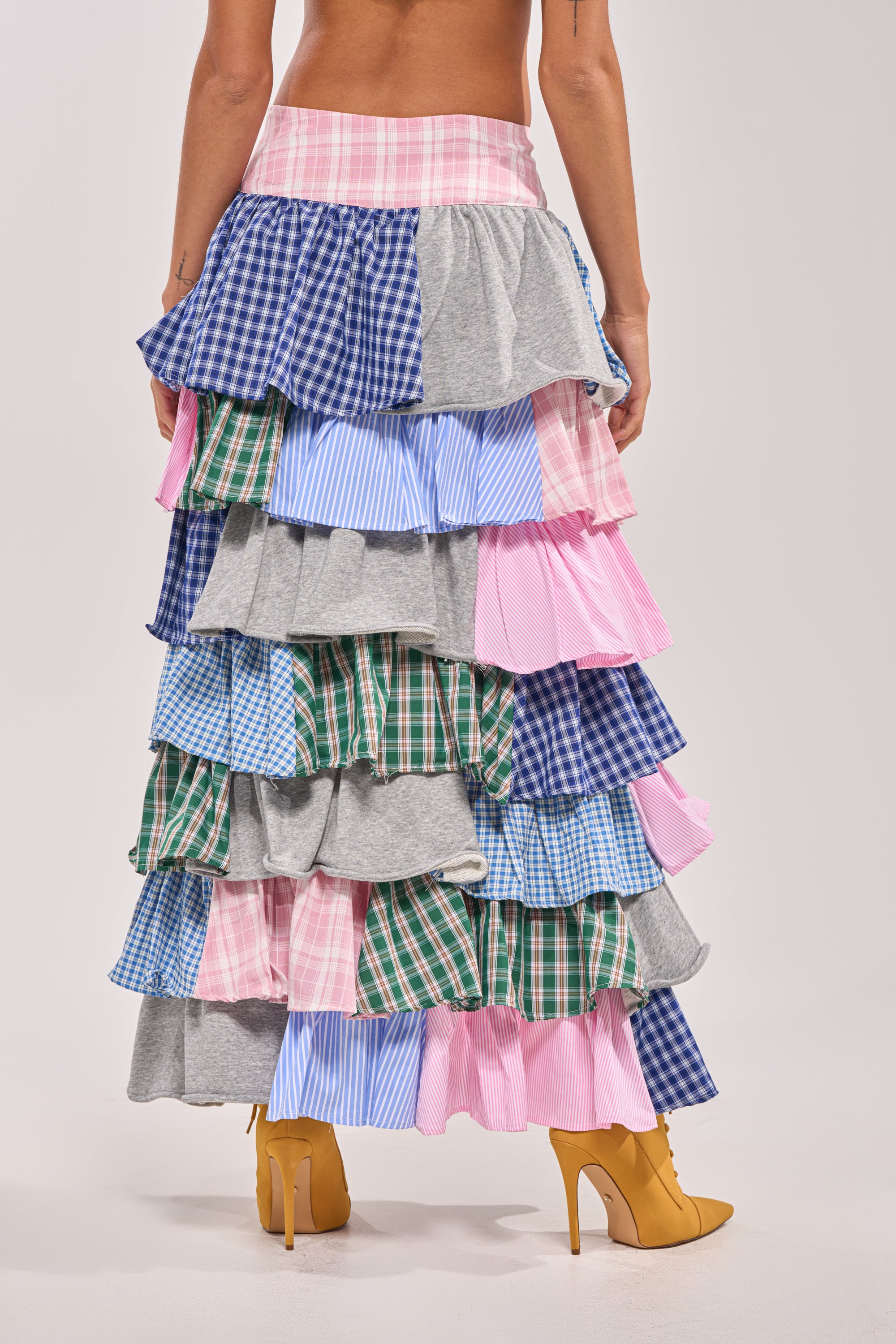 Someone wears the MIXED EMOTIONS PLAID HIGH LOW SKIRT, featuring multi-layered patchwork ruffles in pink, blue, green, and gray. Paired with mustard yellow heeled boots, they face away from the camera. 5