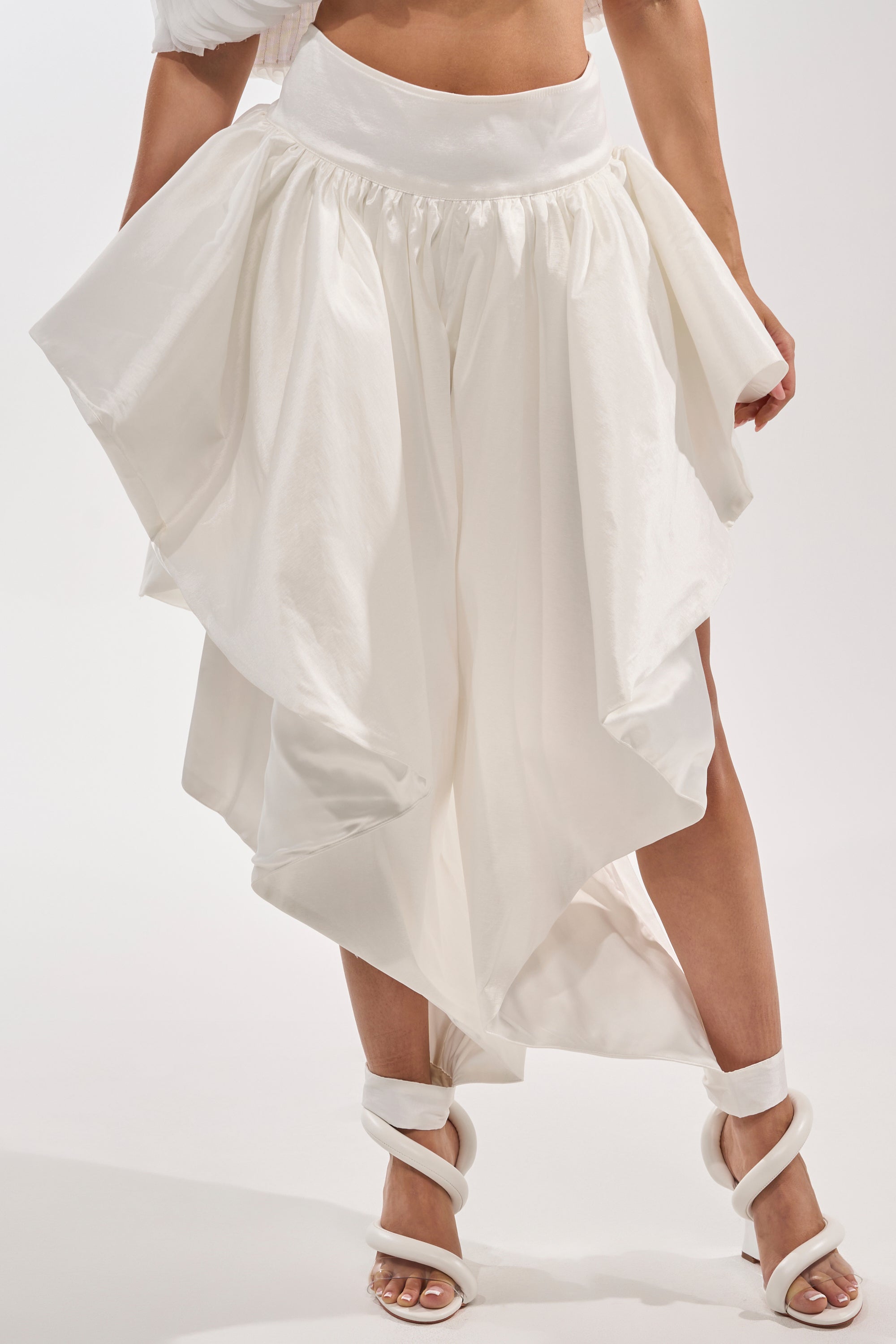 Model showcases the COOL GIRL SKIRT PANT, a white asymmetrical flowing skirt pant with a high waistband, paired with matching strappy heeled sandals. Shot from the waist down against a plain backdrop for an avant-garde look. 1