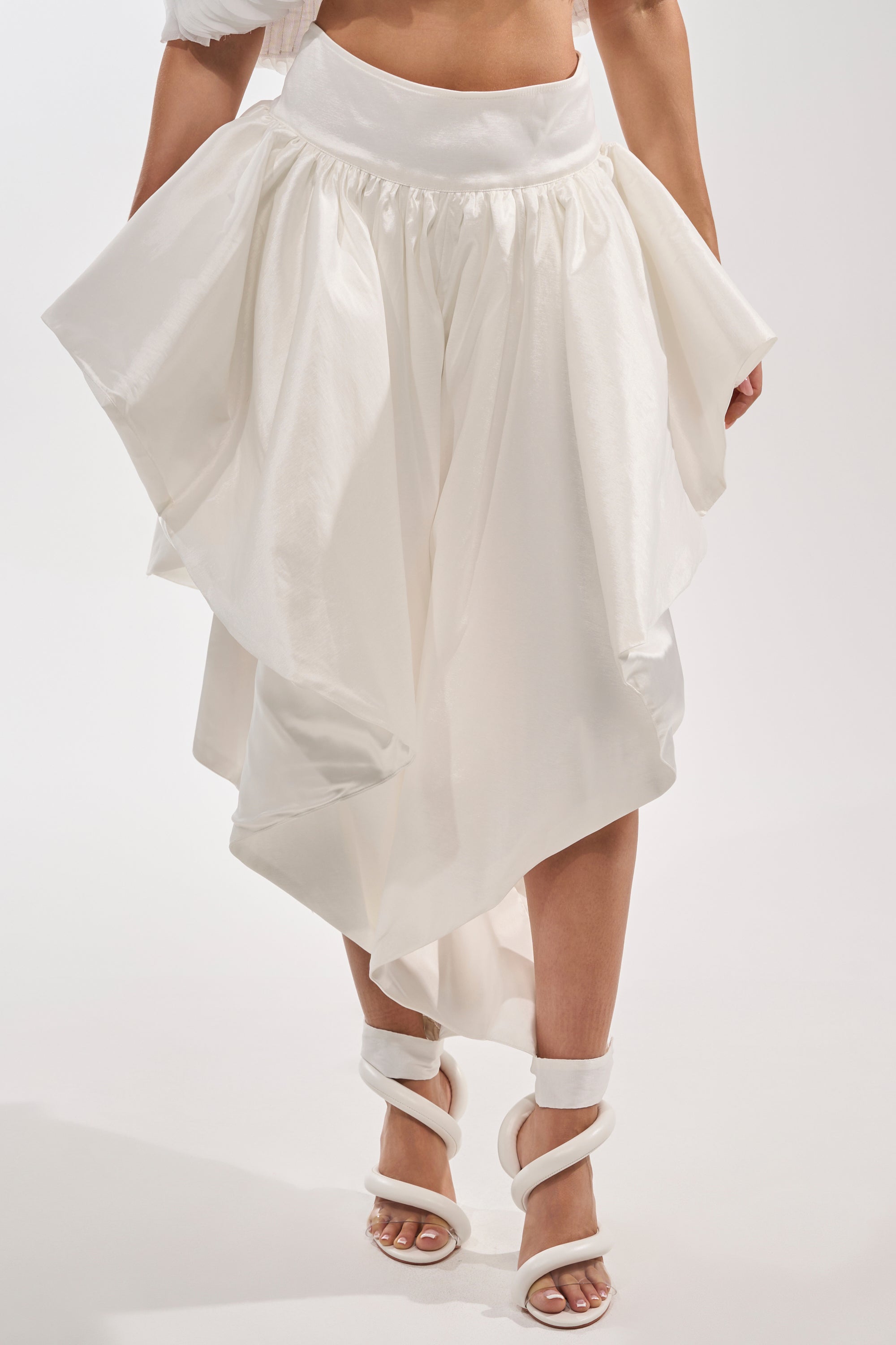 A person wears the COOL GIRL SKIRT PANT in white, featuring an asymmetrical, high-waisted silhouette with a voluminous eveningwear look, paired with matching white strappy high-heeled sandals against a plain background. 2