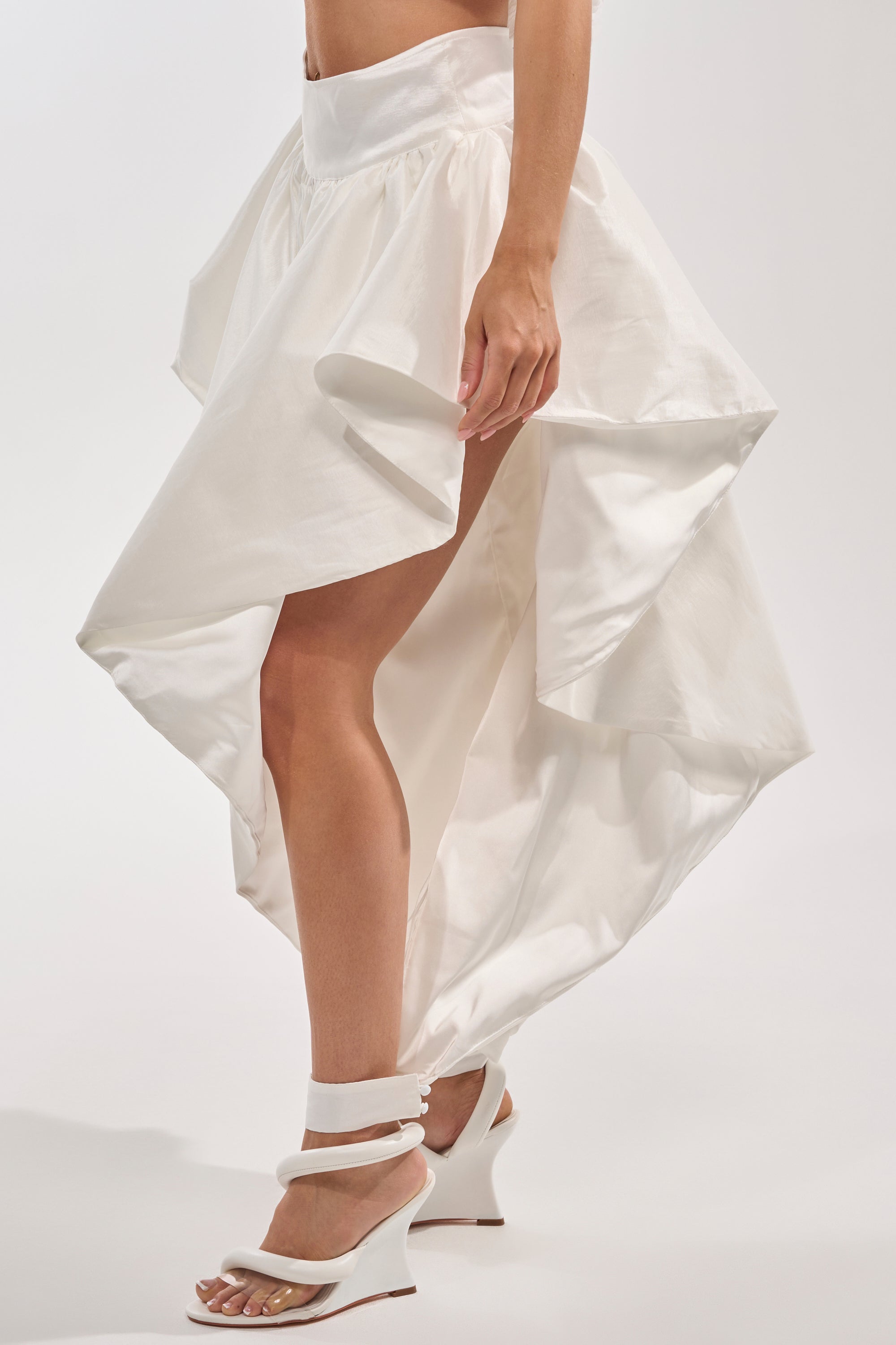 A person wears the COOL GIRL SKIRT PANT, a high-low white skirt with bold, structured folds for an avant-garde look, paired with white heeled sandals against a plain light background. 3