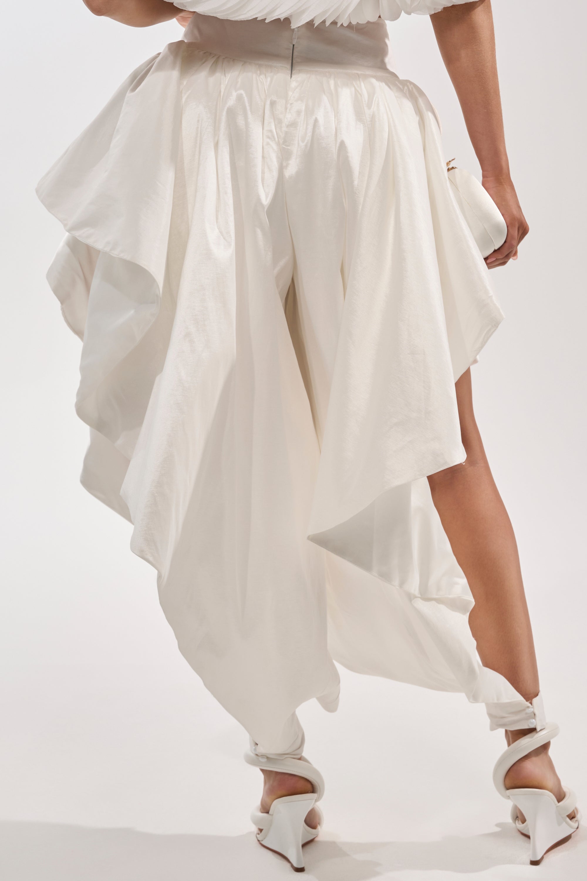 A person wearing the COOL GIRL SKIRT PANT, featuring asymmetrical draped layers and avant-garde styling, with white heeled sandals and a small white clutch, shown from the back against a plain background. 4