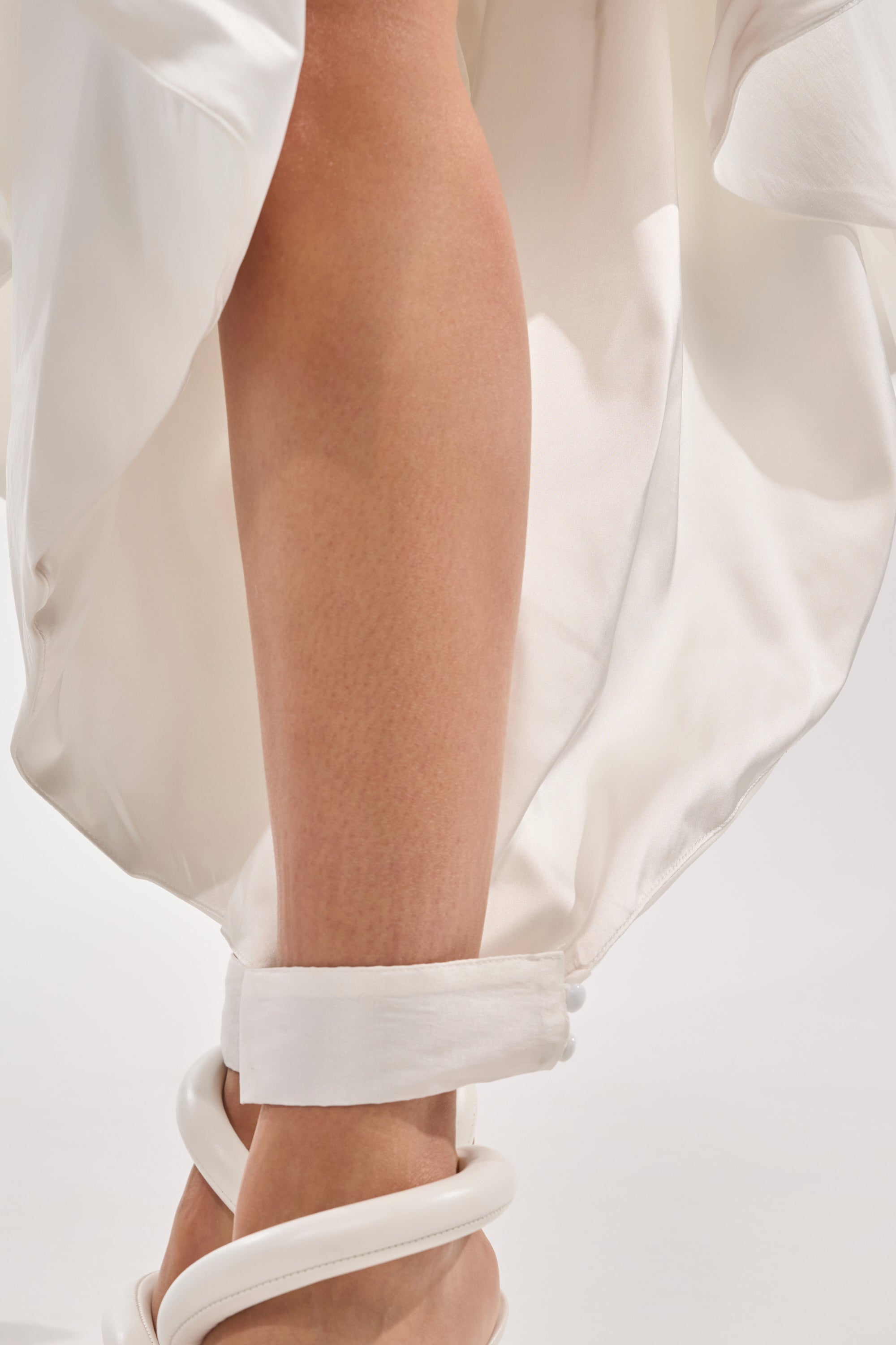 A close-up of a person’s lower leg in the COOL GIRL SKIRT PANT and white heeled sandals with sculptural straps, capturing avant-garde style against a plain, light background. 6