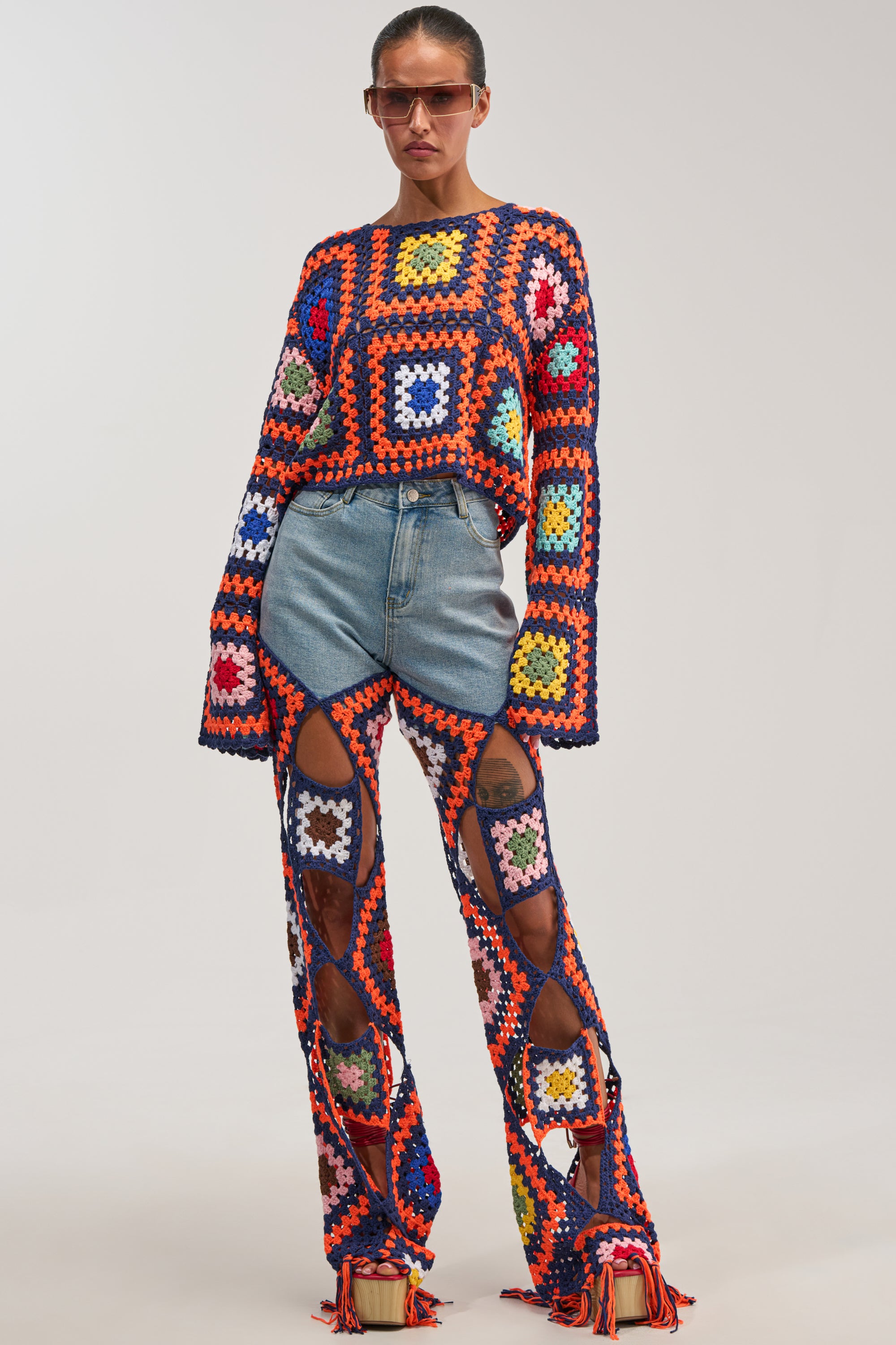 Wearing the MIXED FEELING CROCHET DENIM—a vibrant crocheted sweater with granny square details and crochet denim pants layered over denim shorts—paired with sunglasses and platform sandals, this individual stands confidently against a plain background, perfectly capturing the spirit of festival fashion.