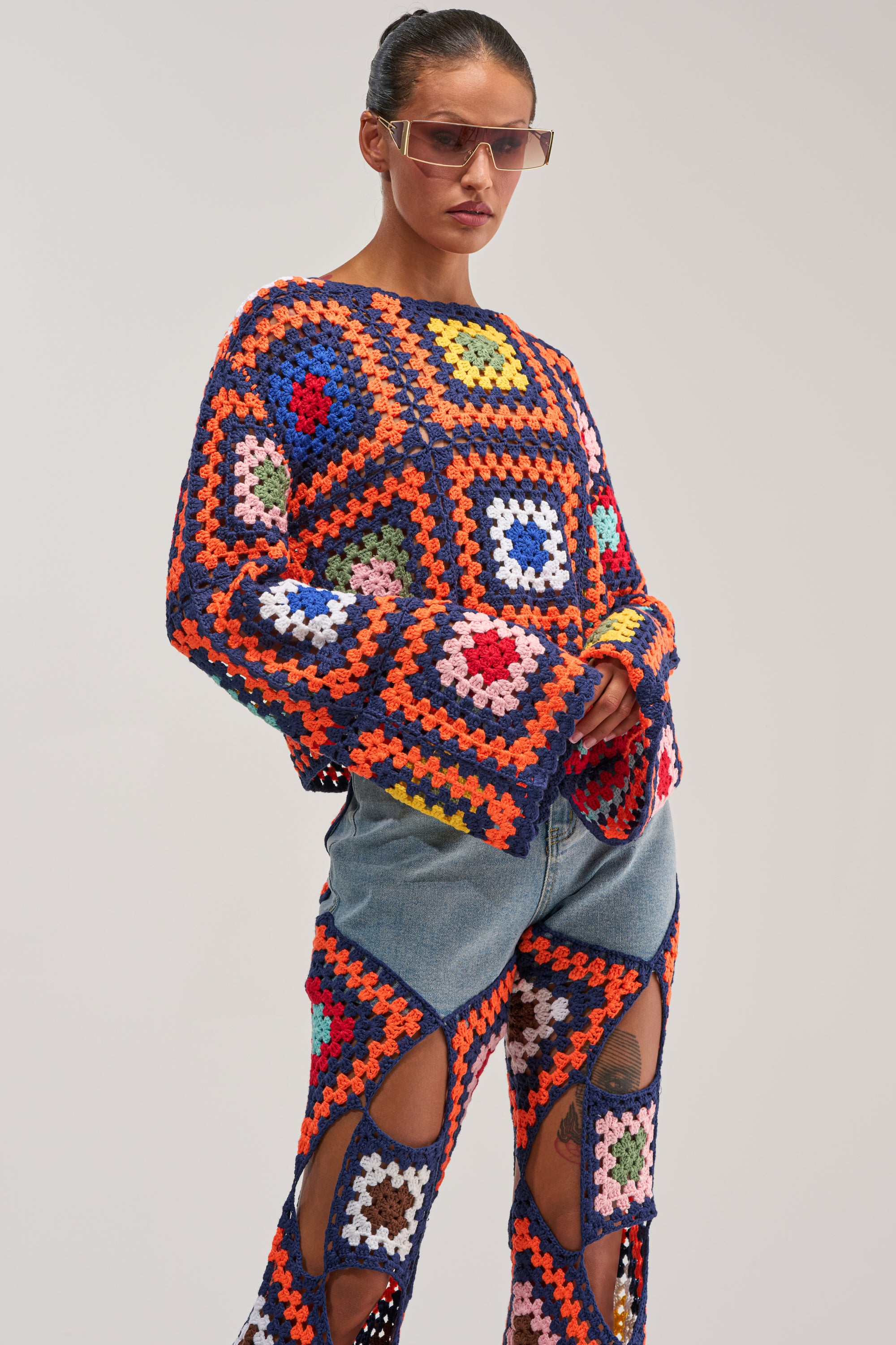 Wearing the MIXED FEELING CROCHET SWEATER and matching patchwork pants layered over denim, a person poses confidently against a plain background, completing the look with pink sunglasses.