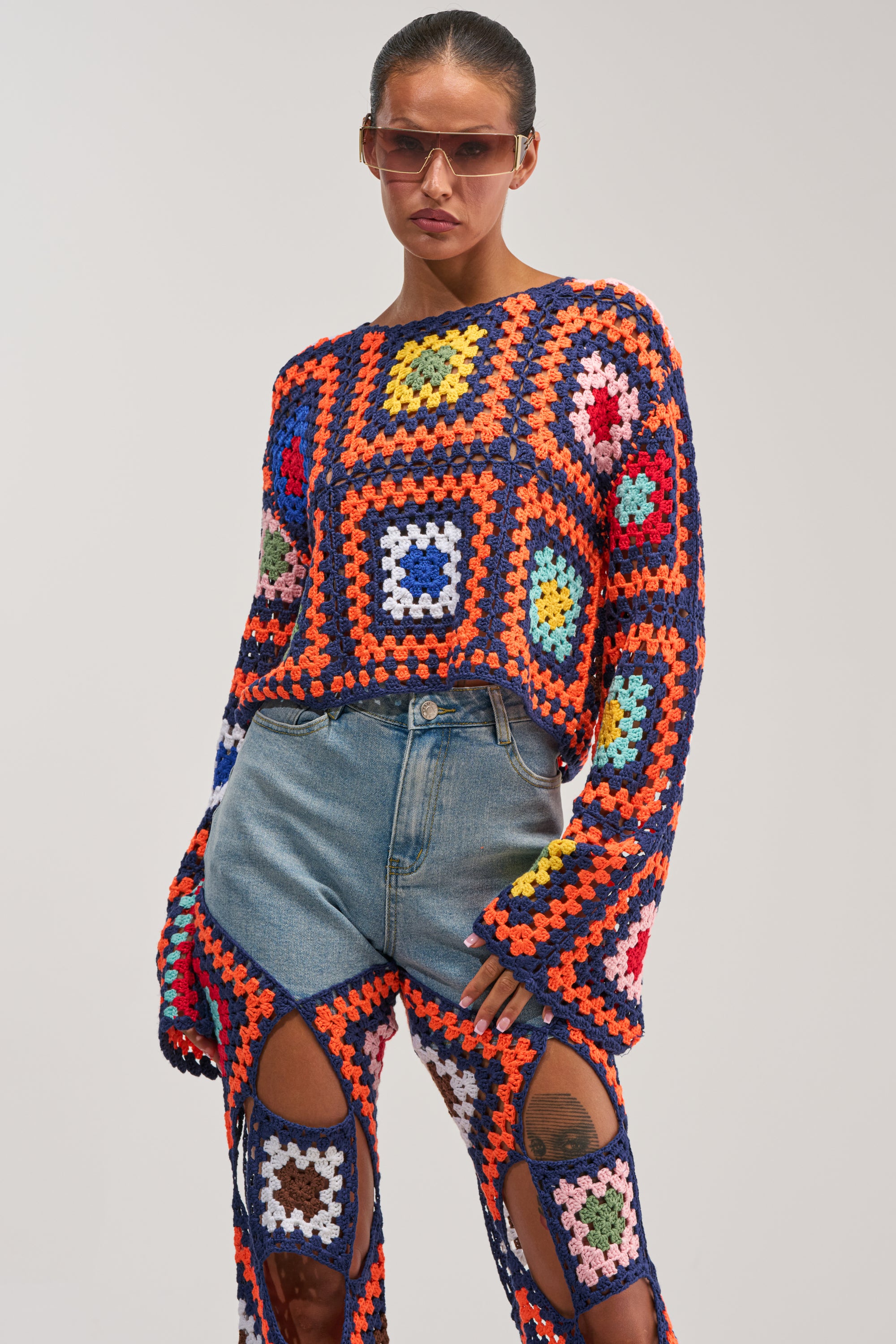 A person wearing the MIXED FEELING CROCHET SWEATER—a vibrant, colorful patchwork crochet sweater with a granny square pattern, styled as a long-sleeve top with matching pants featuring playful cut-out sections—stands confidently against a plain background, sunglasses completing the bold look.