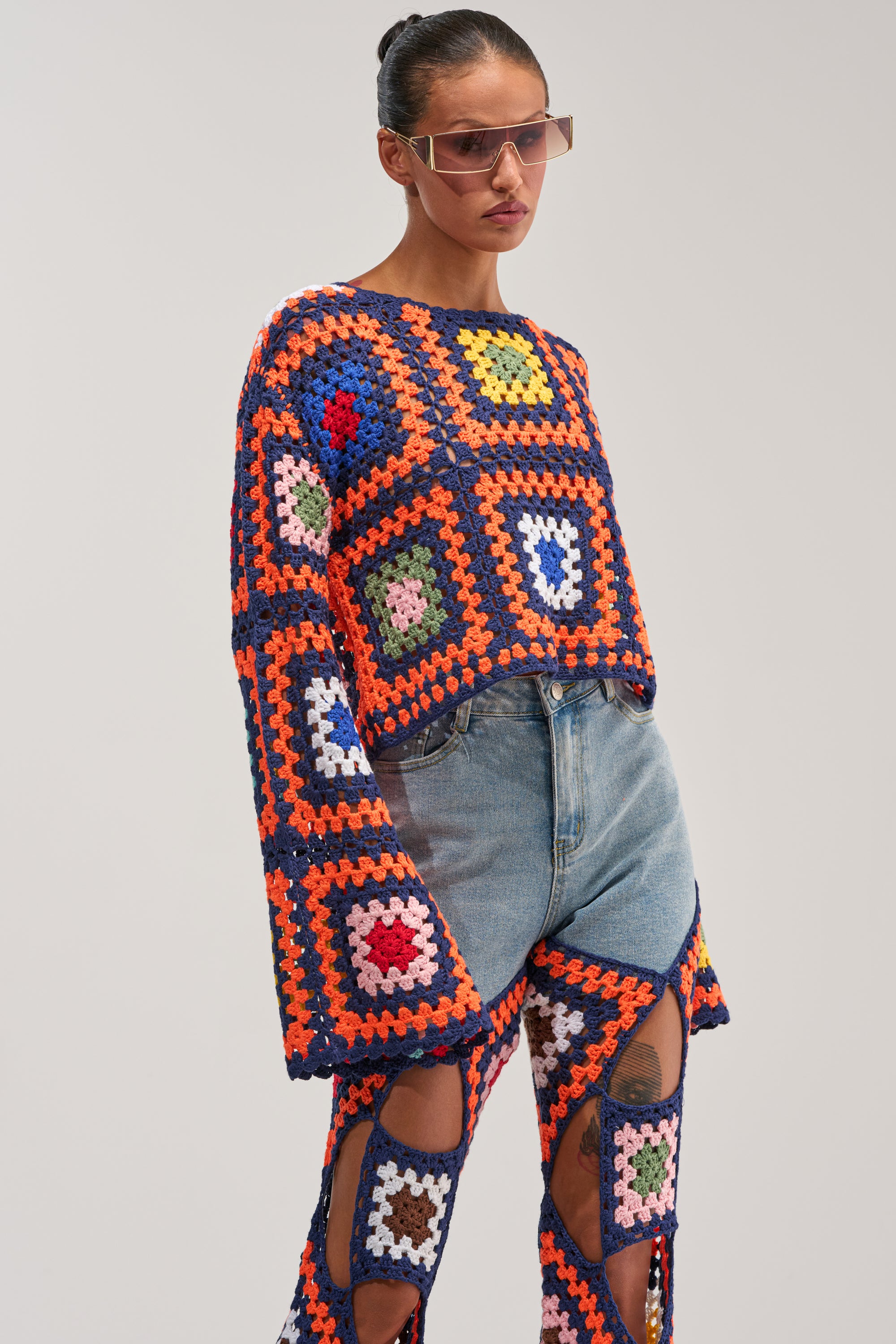 A person wearing sunglasses and the MIXED FEELING CROCHET SWEATER with matching patchwork crochet pants over denim shorts stands against a plain background.