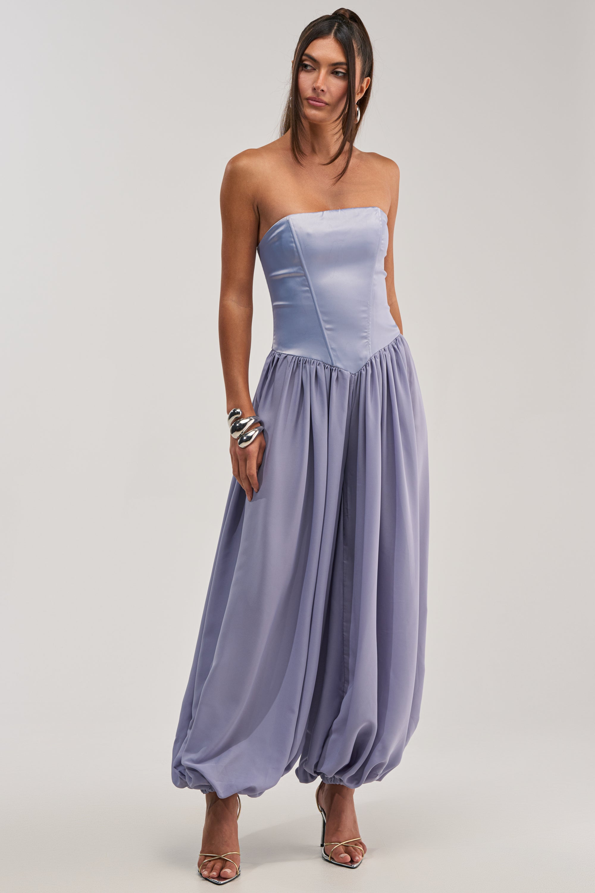 A woman wears the READ MY MIND STRAPLESS JUMPSUIT IN BLUE, designed with a fitted bodice and wide ankle-gathered pant legs. Paired with heels and jewelry against a plain background, it's an elegant choice for formal occasions.
