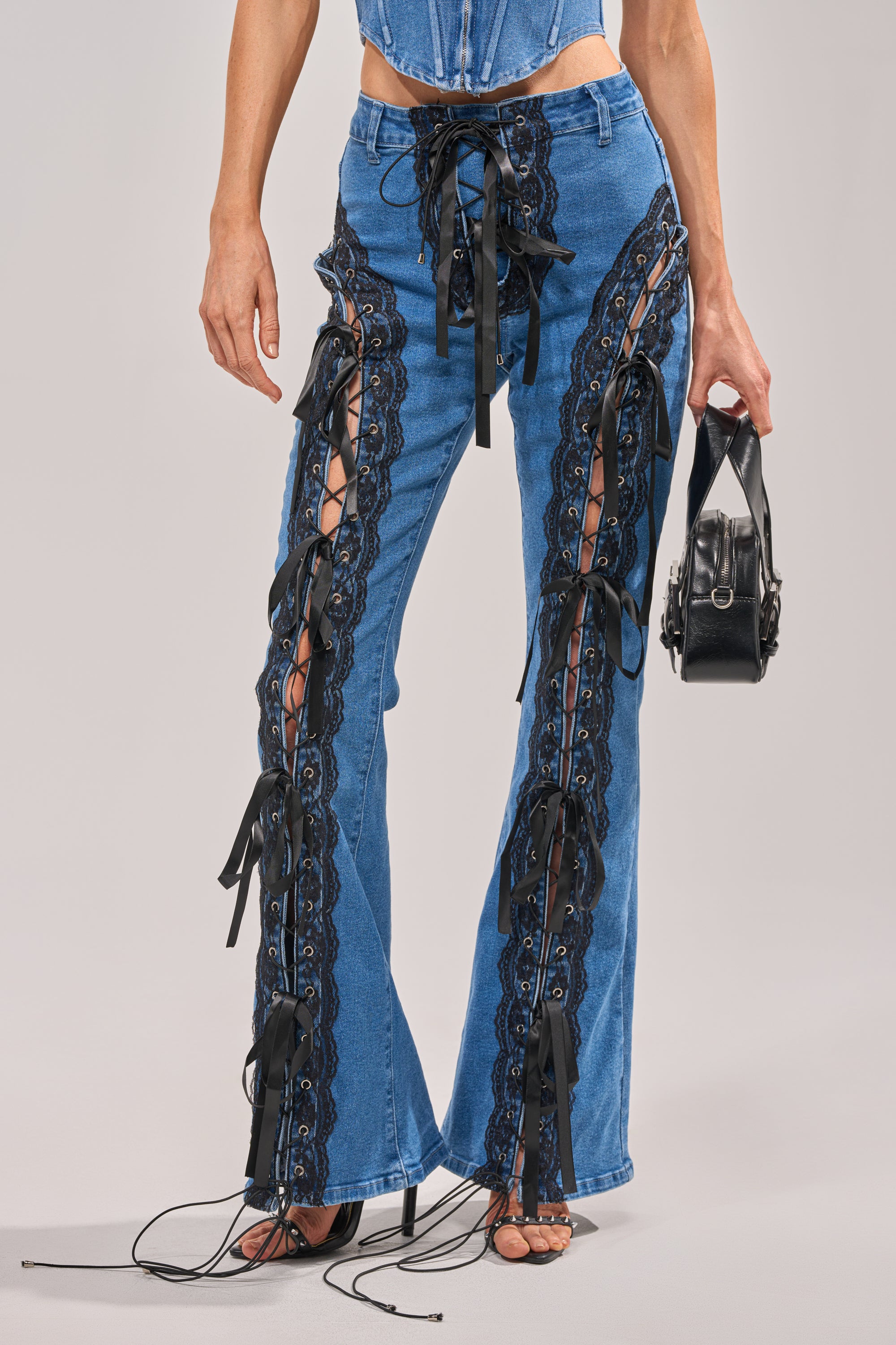 Someone wearing the LACED UP DENIM flared pants, featuring black lace-up details, side cutouts, and multiple black ribbon ties for an adjustable bungee cord effect, holds a black handbag. 0