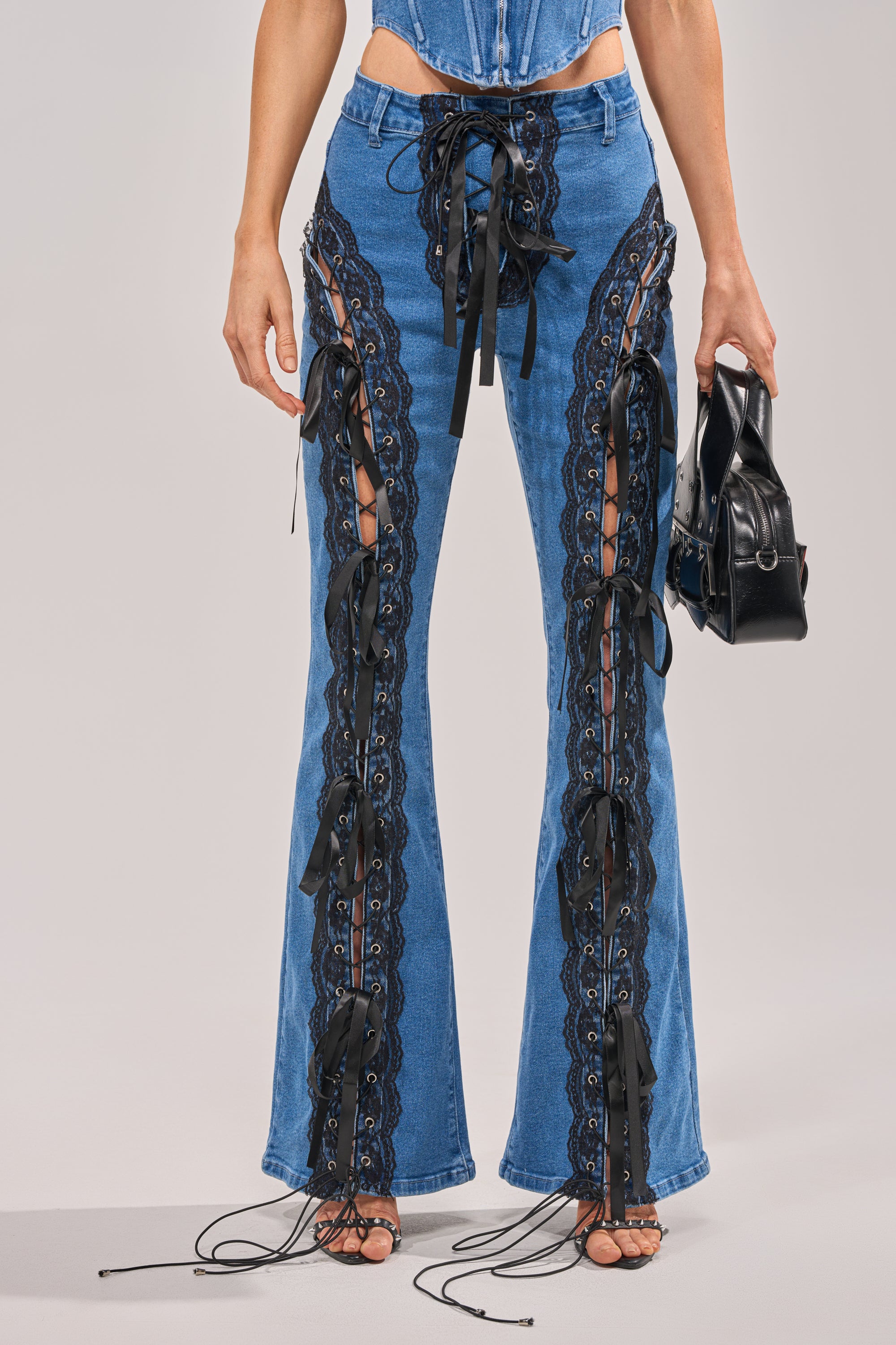 Someone styled in LACED UP DENIM, blue jeans with a customizable fit and black ribbon ties along side cutouts, paired with a matching top and carrying a small black handbag. 1