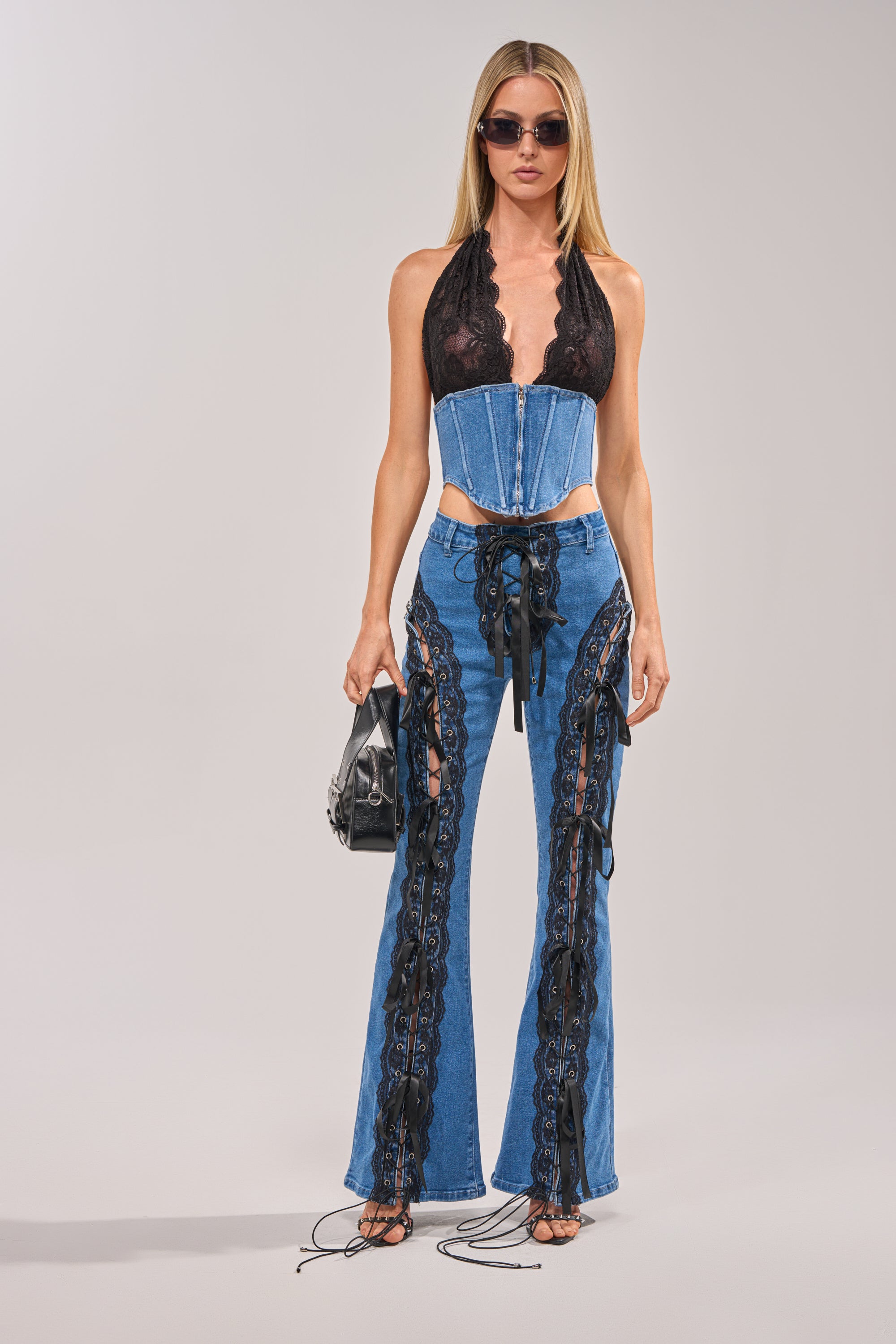 A woman with straight blonde hair stands holding a black handbag, wearing LACED UP DENIM jeans with blue lace trim and black lace details, a black lace halter top, sunglasses, and strappy sandals. 4