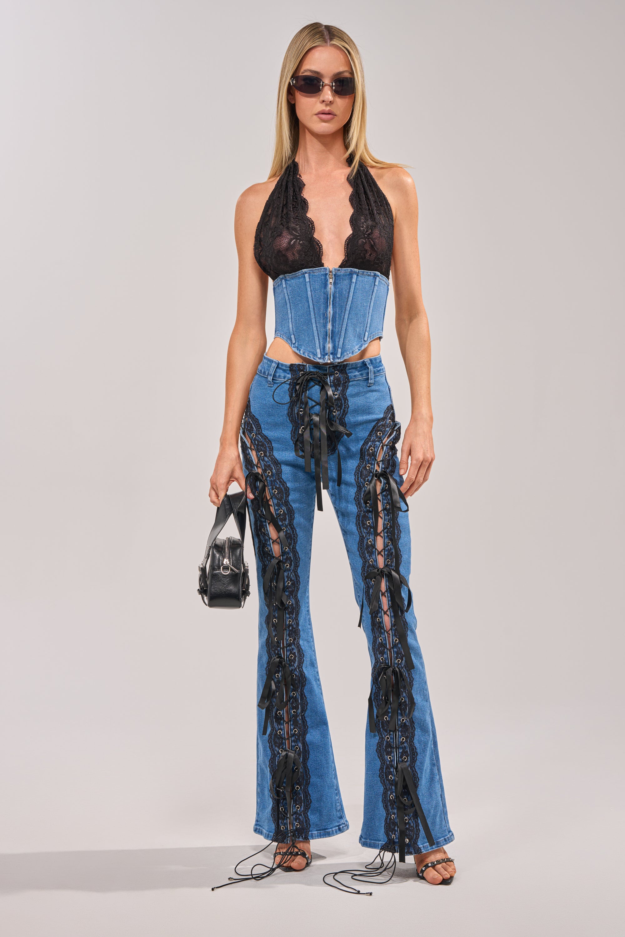 A woman stands confidently against a plain background, wearing the LACED UP DENIM set with a black lace halter top, denim corset, and flared pants featuring black lace-up details, accessorized with sunglasses and a small black handbag. 5