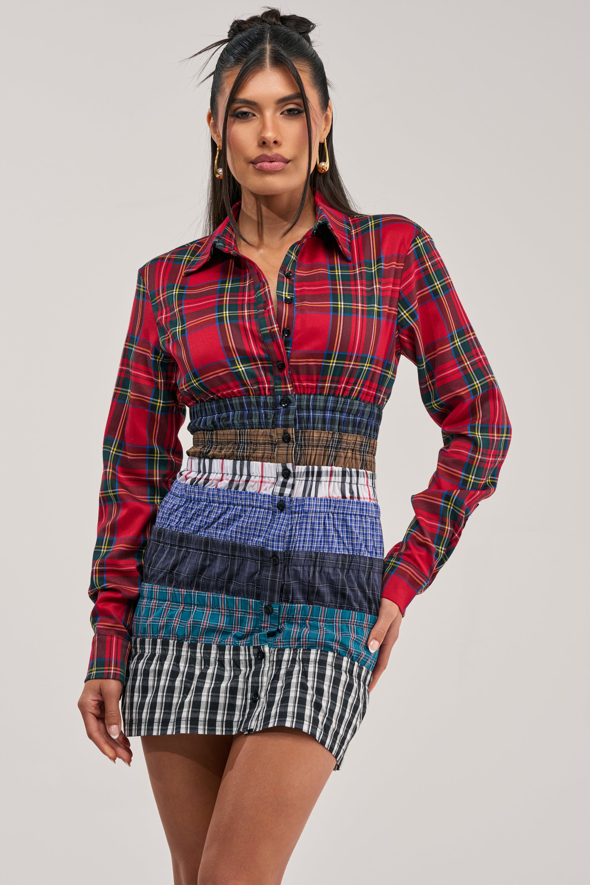 A woman models the LAYERS TO MY LOVE FLANNEL MINI DRESS, featuring a street style design with a red plaid top and a skirt composed of horizontal stripes in various plaid and check patterns, standing against a plain light background.