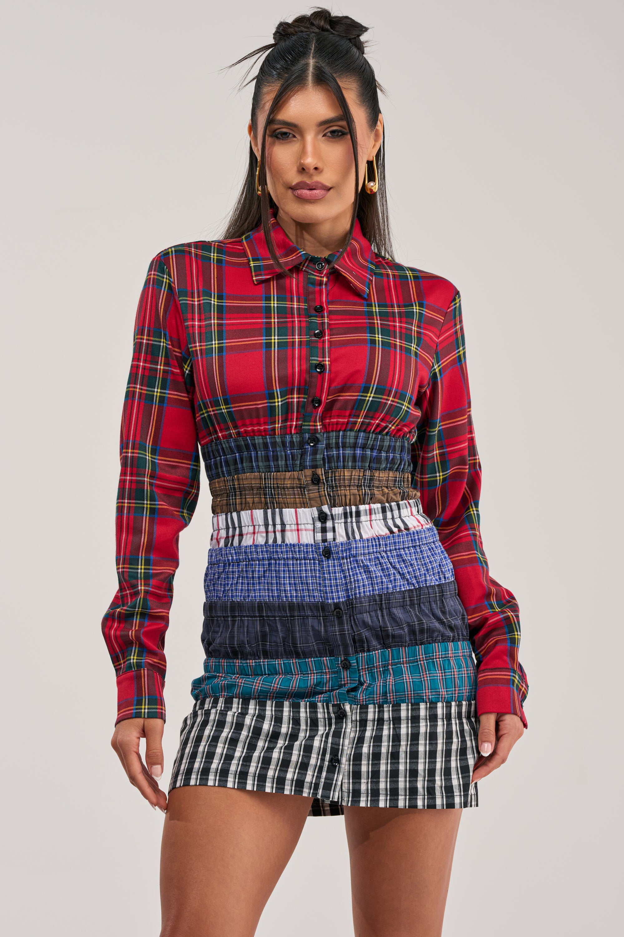 A woman exhibits statement street-style flair in the LAYERS TO MY LOVE FLANNEL MINI DRESS, featuring a red plaid top and a layered skirt of multicolored plaid and striped fabric, as she poses against a plain background.