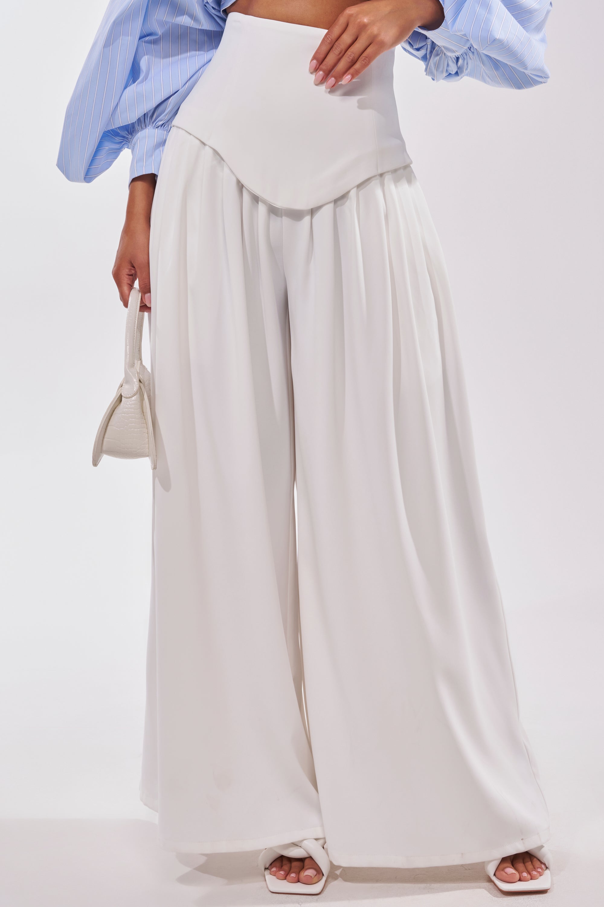 A person is seen from the waist down wearing GALAXY TROUSER high-rise white wide-leg trousers, a light blue puff-sleeve top, white heeled sandals, and holding a small white handbag. 0