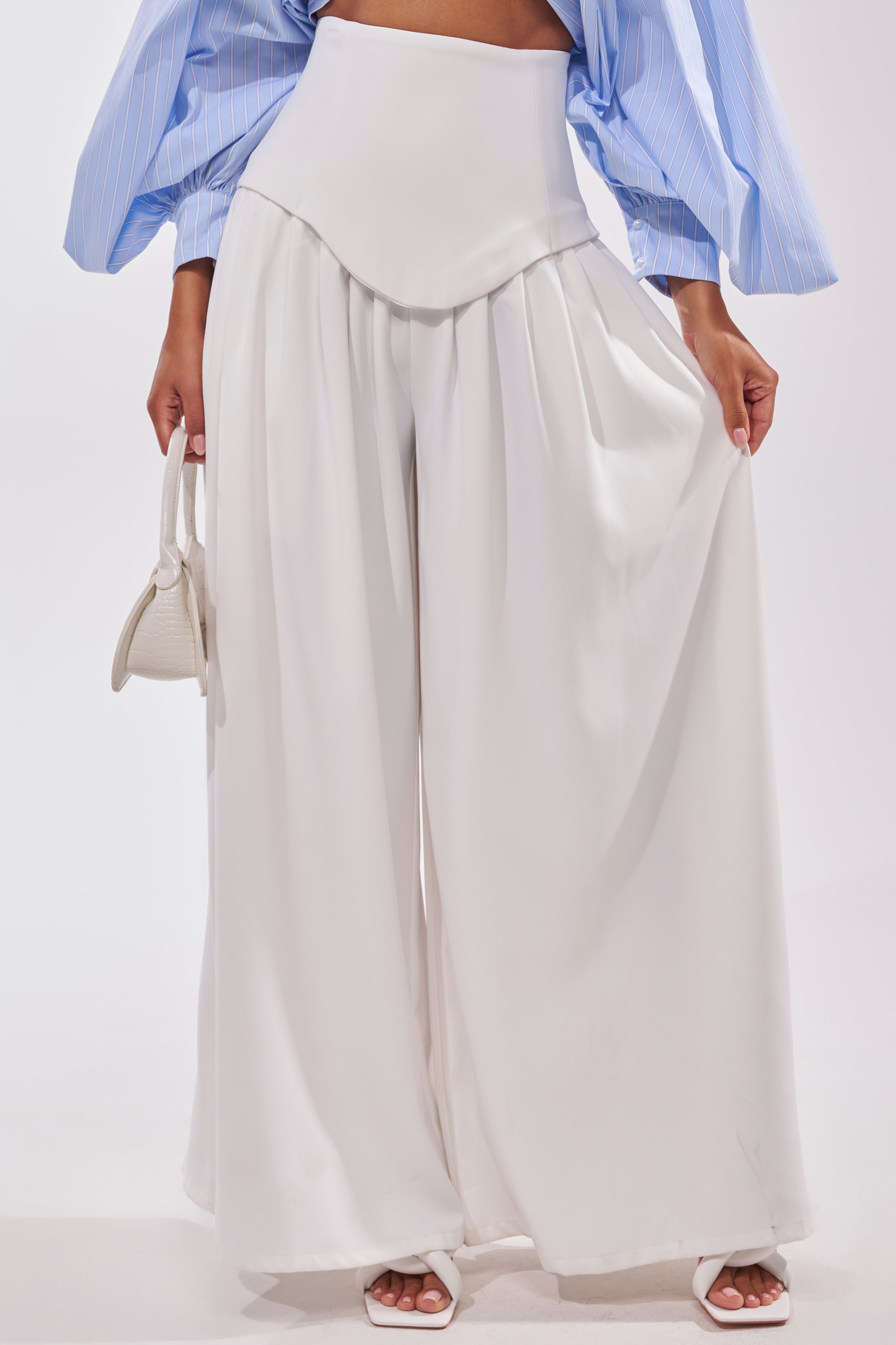 A person is shown from the waist down wearing GALAXY TROUSER—high-rise, wide-leg, white trousers—paired with a light blue long-sleeve puffed blouse, white sandals, and holding a small white handbag. 1