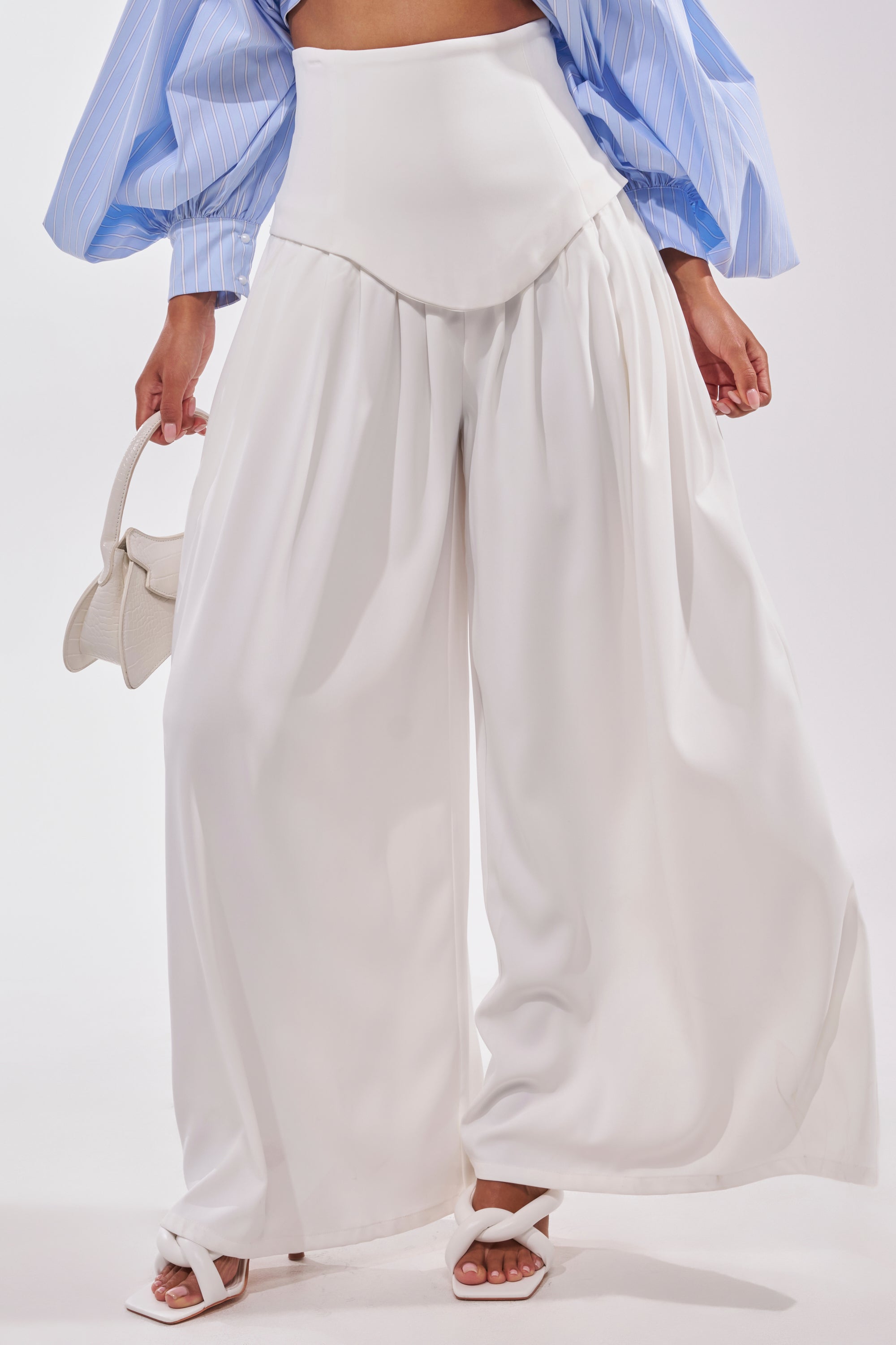 A person wears the GALAXY TROUSER—white, high-rise, wide-leg trousers—with a blue crop top featuring puffy sleeves, white heeled sandals, and carries a small white handbag. The background is plain white. 2