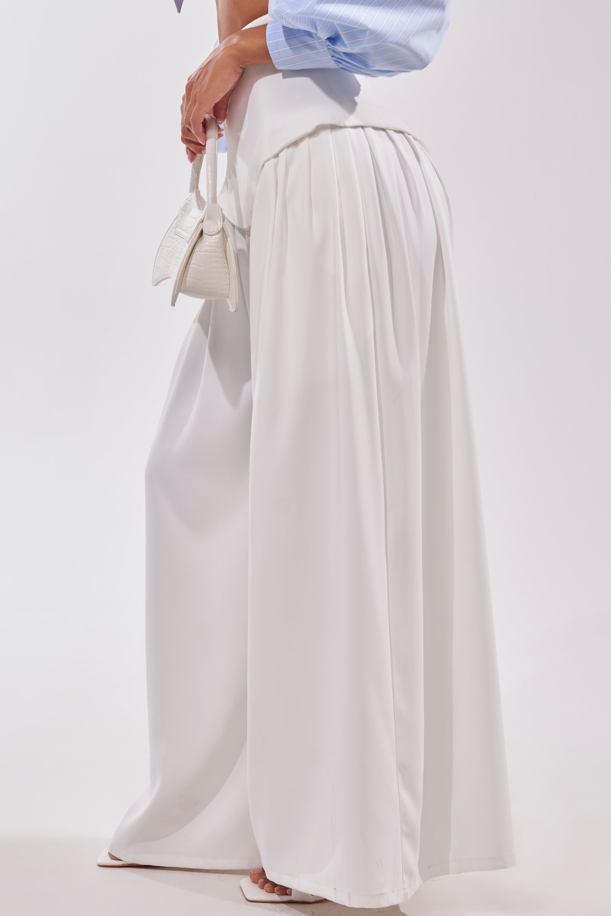 A person displays the GALAXY TROUSER, a high-rise waist, white wide-leg trouser, paired with a light blue long-sleeve shirt and holding a small white handbag. Only the lower half is shown against a plain background. 3