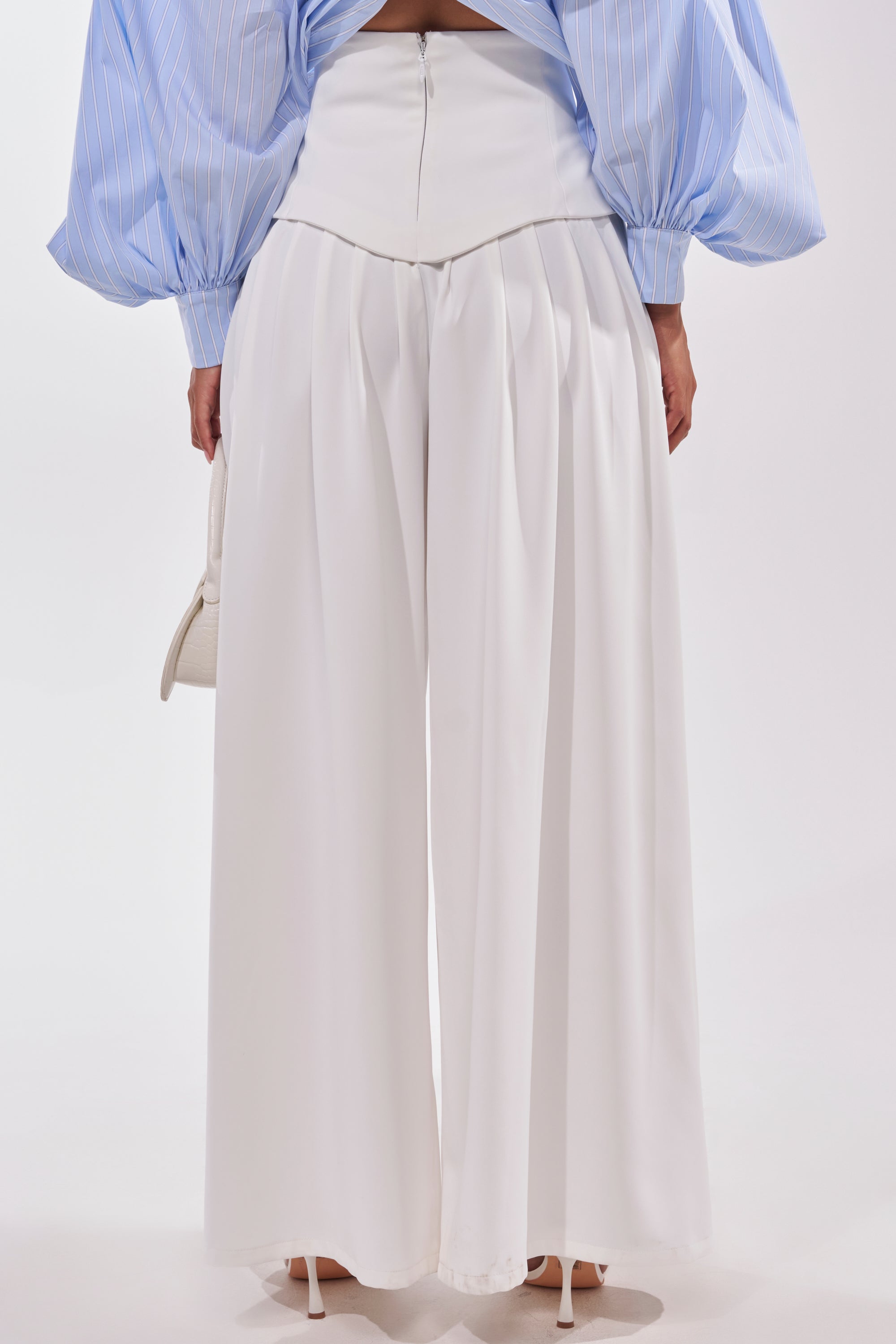 Wearing the GALAXY TROUSER—flowy, high-rise white wide-leg pants with pleats—a person pairs them with a blue and white pinstripe blouse with voluminous sleeves and white heels, standing against a plain background. 4