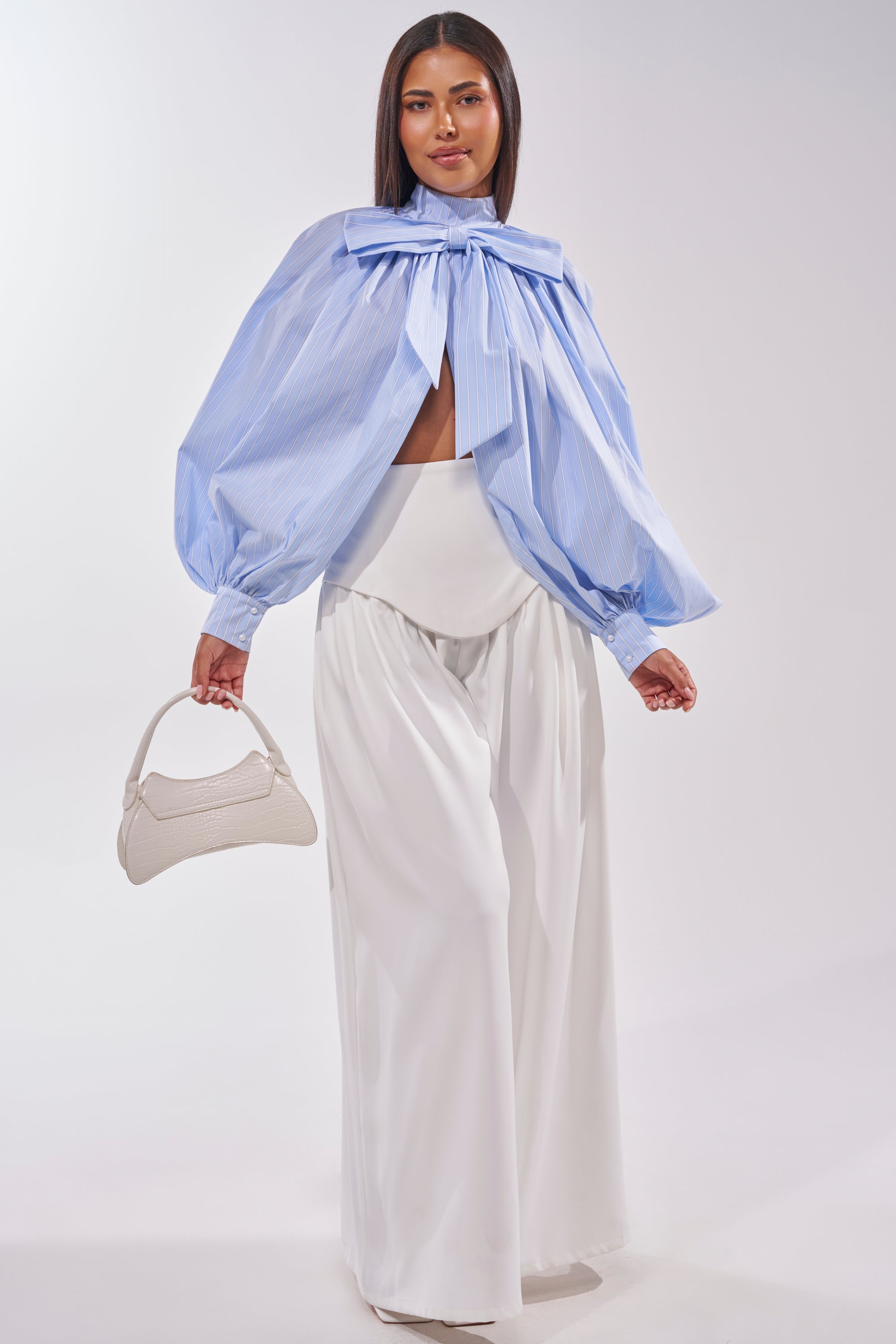 A woman poses against a plain backdrop wearing the GALAXY TROUSER—white, high-rise wide-leg pants—paired with a light blue long-sleeve top with a large neck bow and voluminous sleeves, and holding a small white handbag. 5