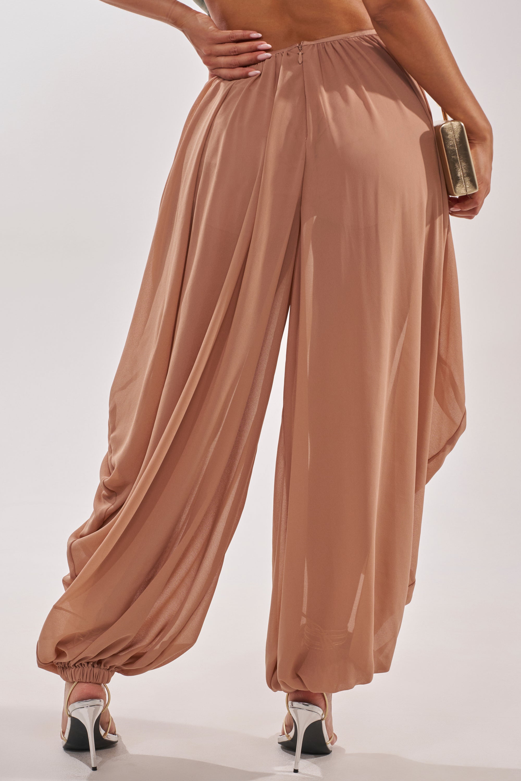A person is seen from the back wearing the BOUNCIN AROUND BALLOON PANT in peach with gathered ankles, paired with white high-heeled sandals and holding a gold clutch against a plain background. 4