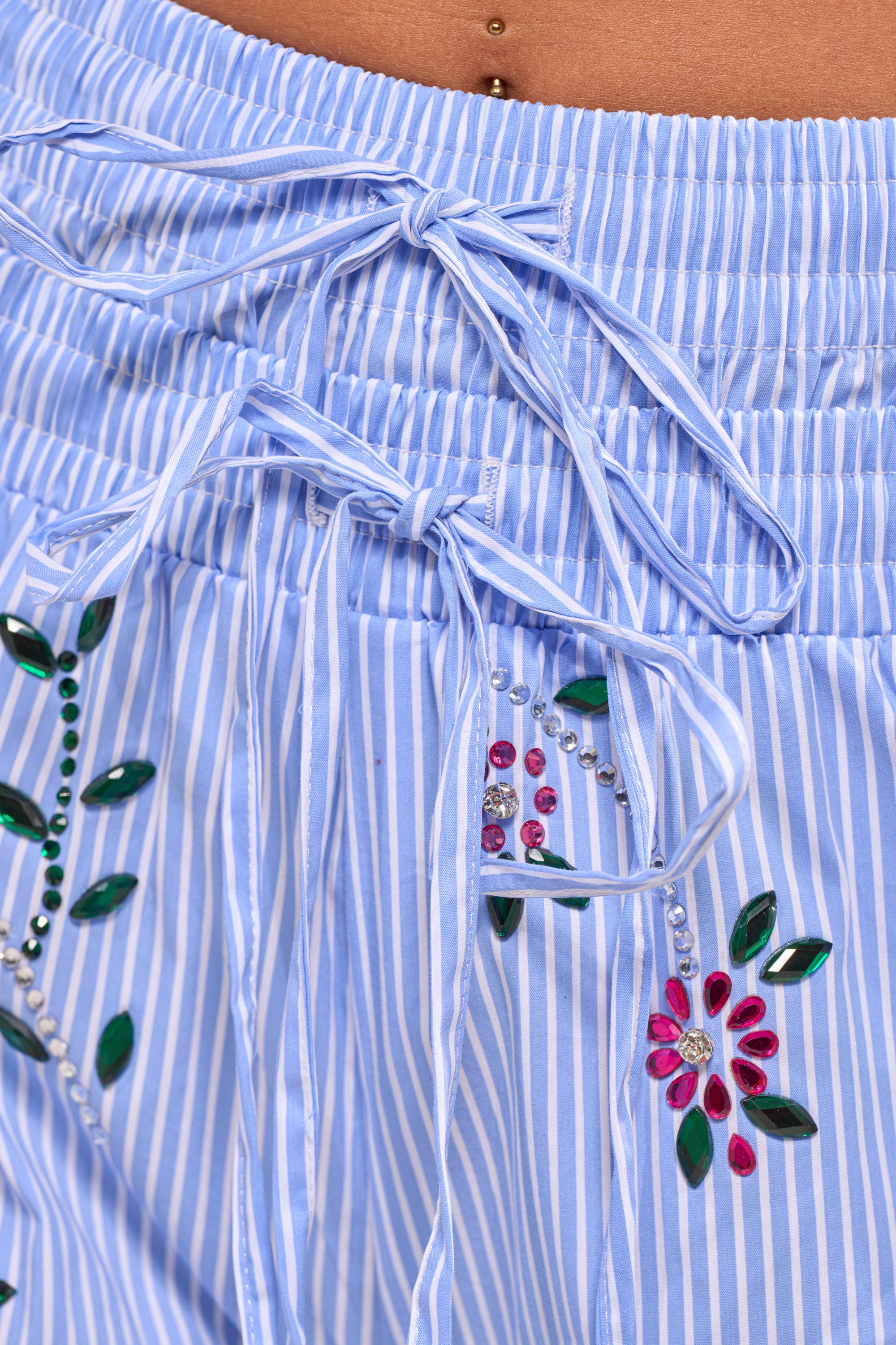 Close-up of the JUNIE POPLIN RHINESTONE DETAIL SHORT, featuring blue and white vertical stripes, floral embroidery, rhinestone accents, and double drawstring bows at the waistband. A belly button piercing is visible above the shorts. 7