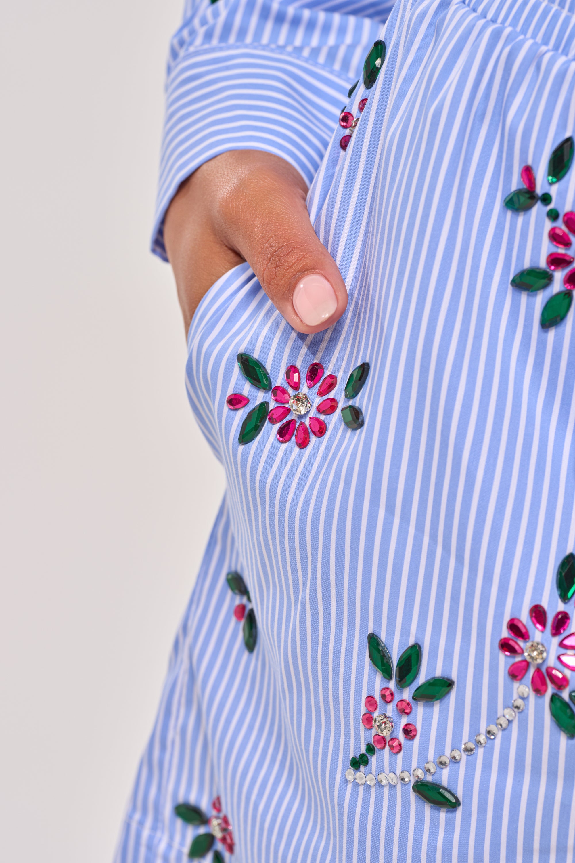 A hand with light pink nail polish rests in the pocket of blue and white striped JUNIE POPLIN RHINESTONE DETAIL SHORTS, adorned with floral embroidery and colorful rhinestone accents. 8